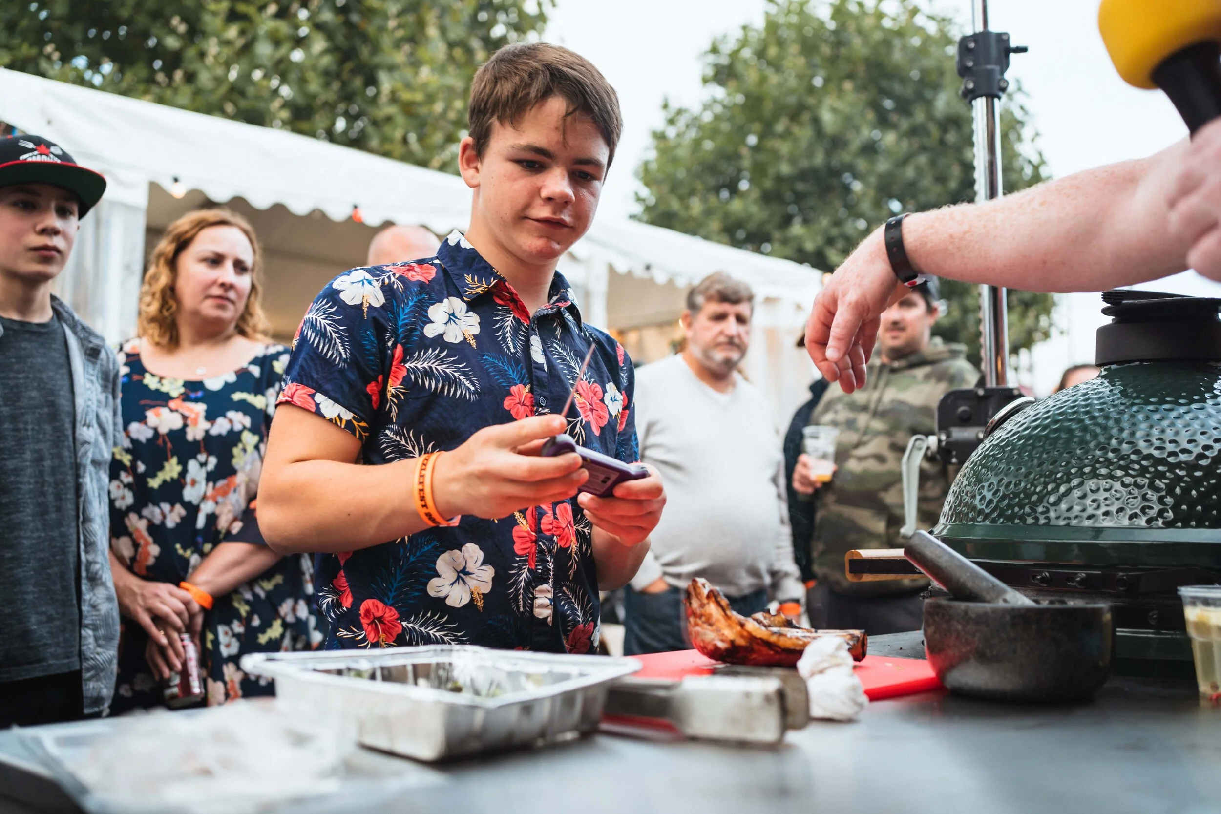 People at an outdoor event with a barbecue, man in floral shirt holding phone, various onlookers, and a piece of grilled meat on a red cutting board in the foreground.