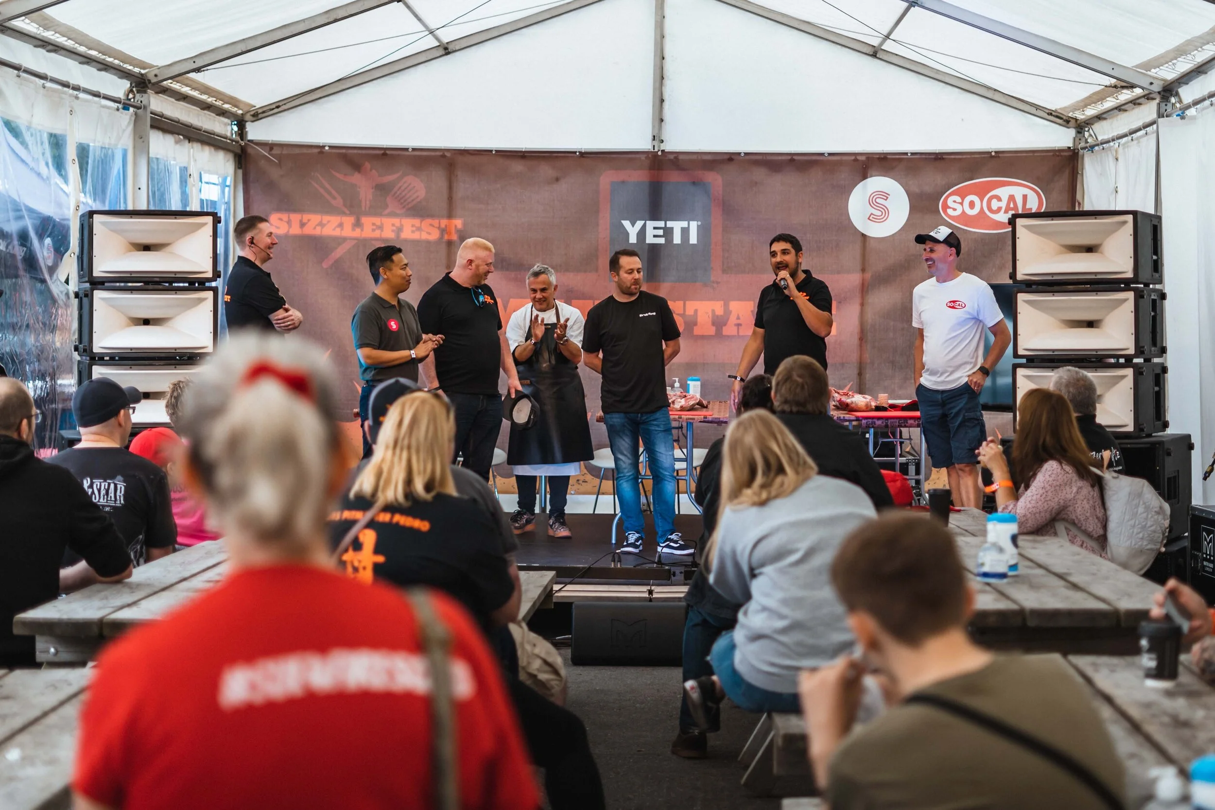 Group of people at a barbecue event, Sizzlefest, seated under a tent with speakers in the background.