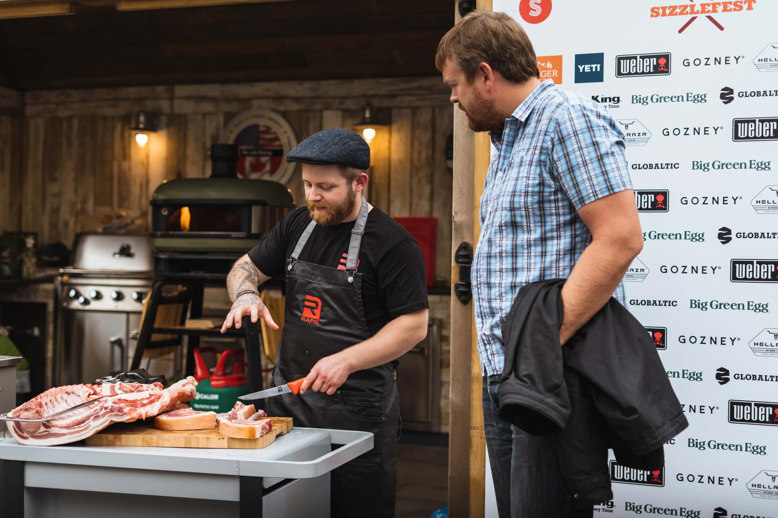 A man in a black apron is cutting meat on a table, observed by another man in a plaid shirt. They're in a cooking space with a pizza oven and grill in the background, with a banner featuring brand logos visible behind them.