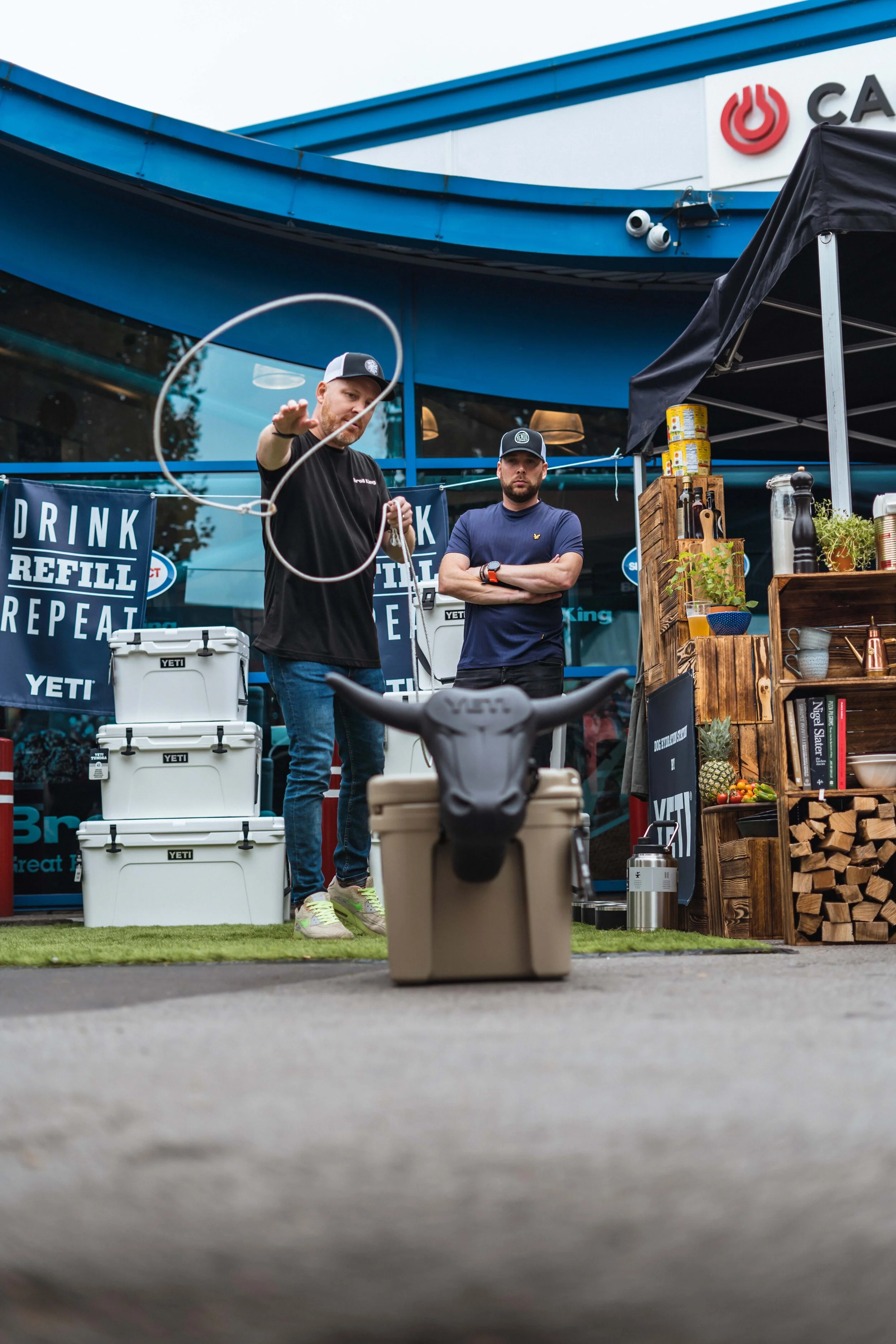 Two people are at an outdoor setup with YETI coolers, where one is attempting to throw a lasso towards a target resembling a bull's head on a cooler. There are wooden crates with products and banners nearby, with a branded canopy overhead. The setup 
