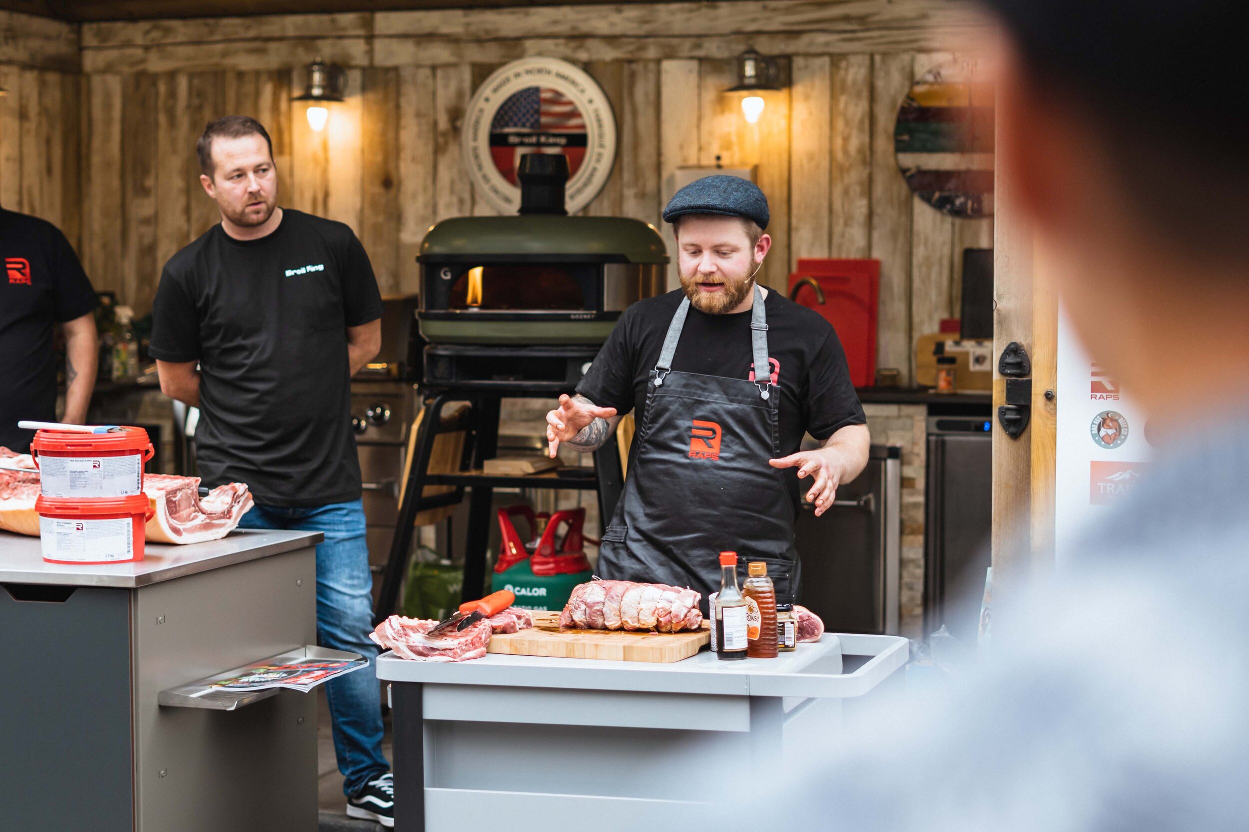 A man in an apron giving a cooking demonstration with meat cuts on a table, surrounded by cooking equipment and people watching. The setting appears to be an outdoor kitchen.