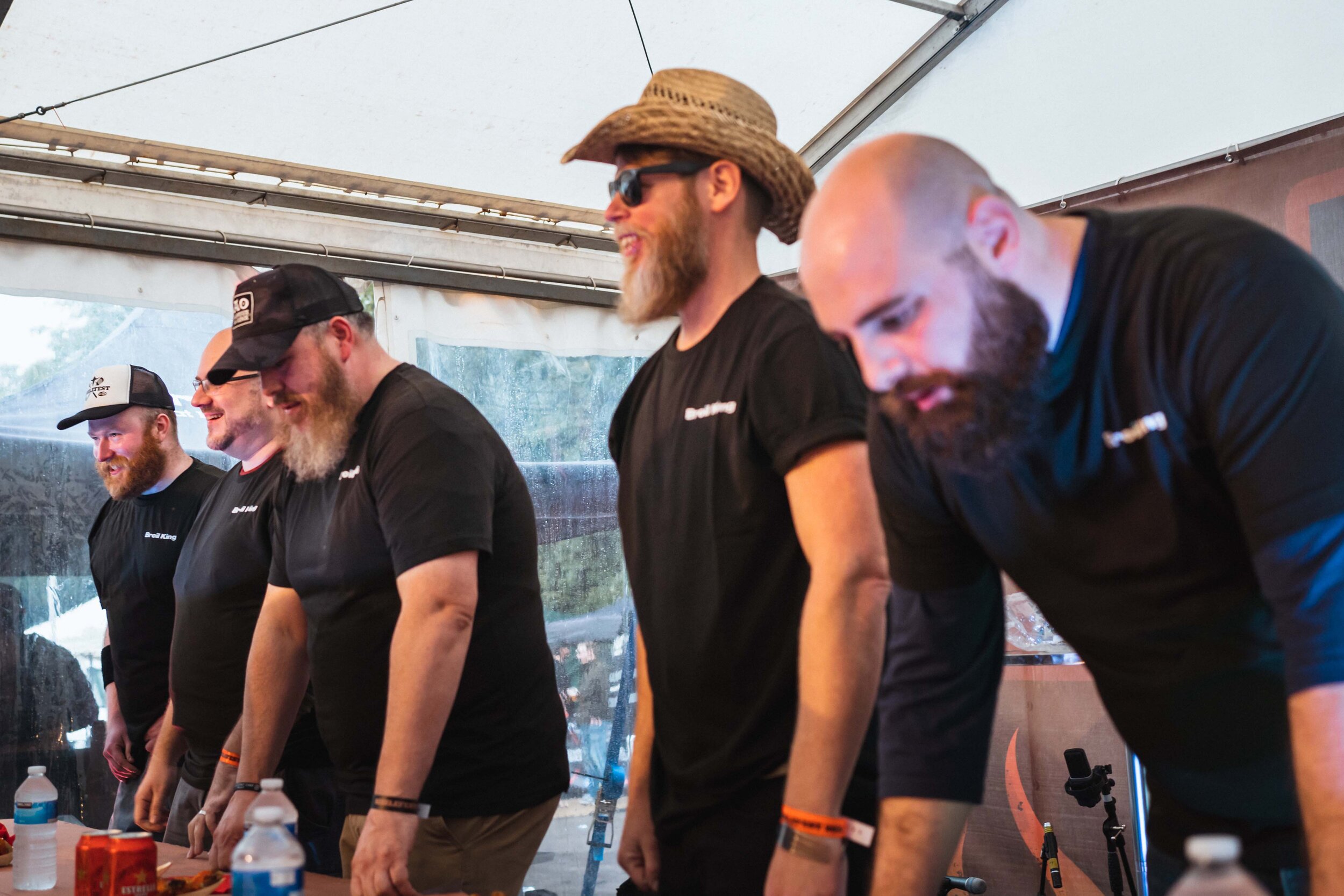 A group of five men dressed in black T-shirts participating in an event, standing at a table under a tent. One wears a straw hat and sunglasses, others wear caps. Water bottles and a red box are on the table.