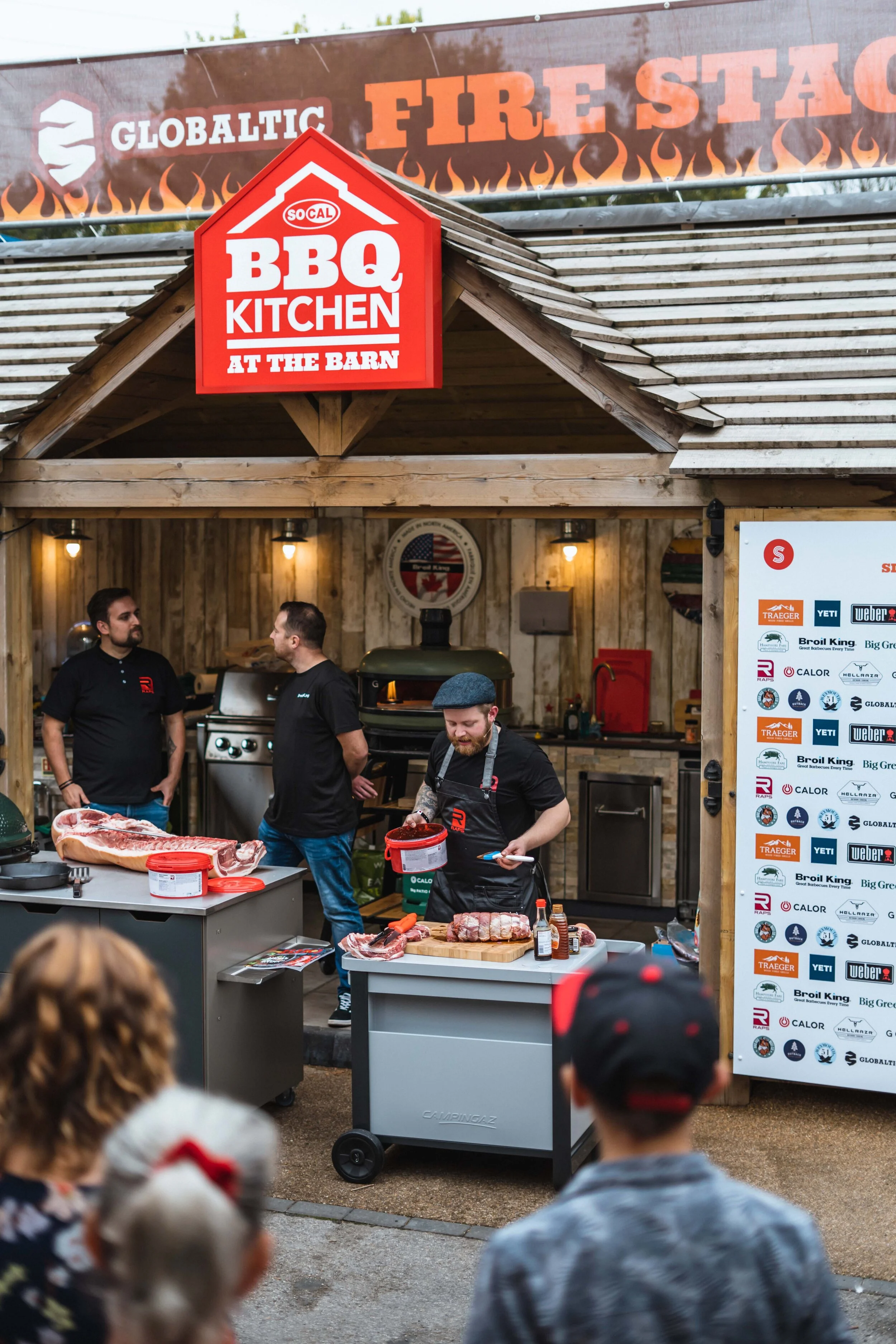 Outdoor BBQ kitchen with chefs preparing meat, branded sign, and audience watching.