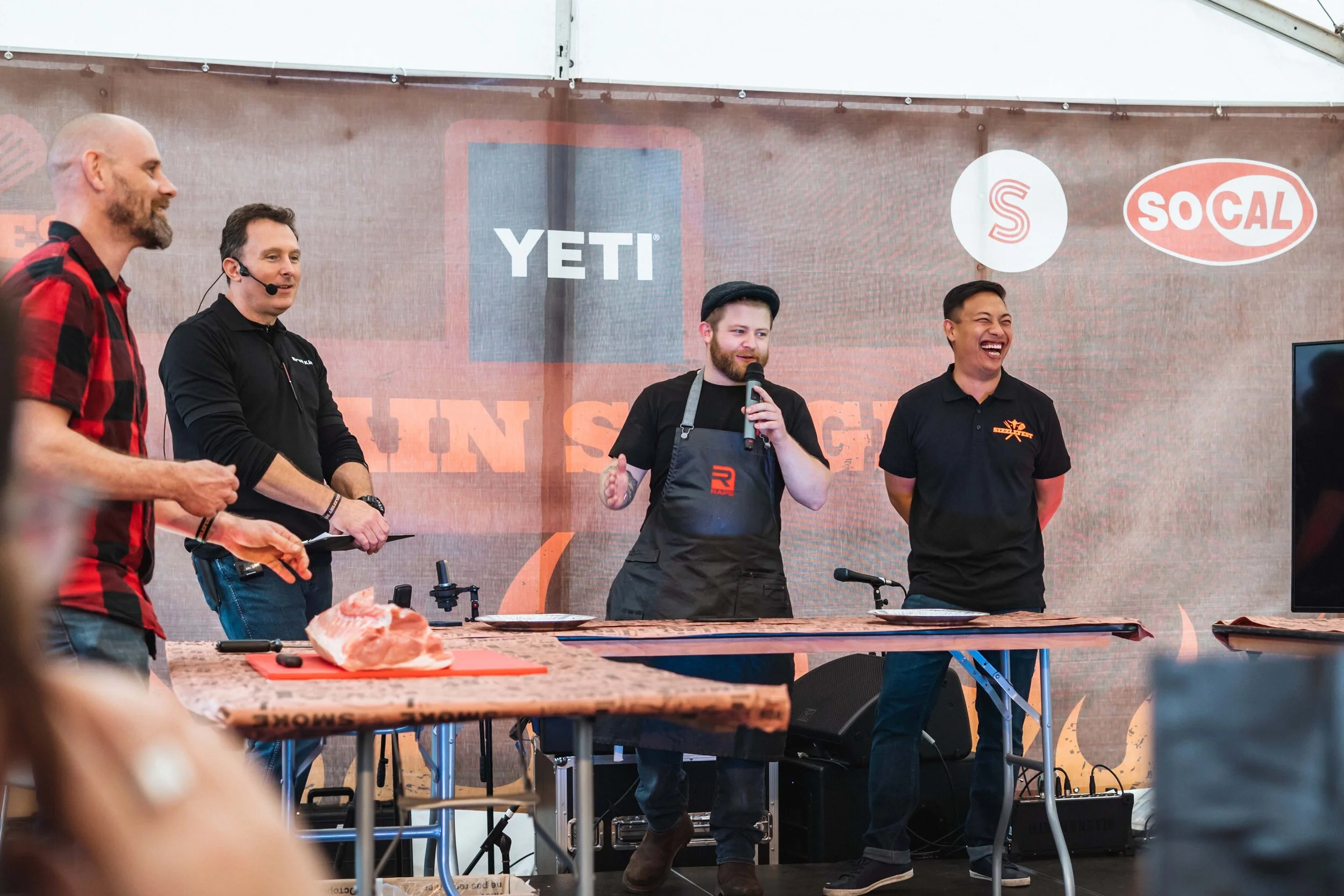 Four men standing around a table during a cooking event, with one holding a microphone. A piece of meat is placed on the table. Banners with brand logos, including YETI, are visible in the background.