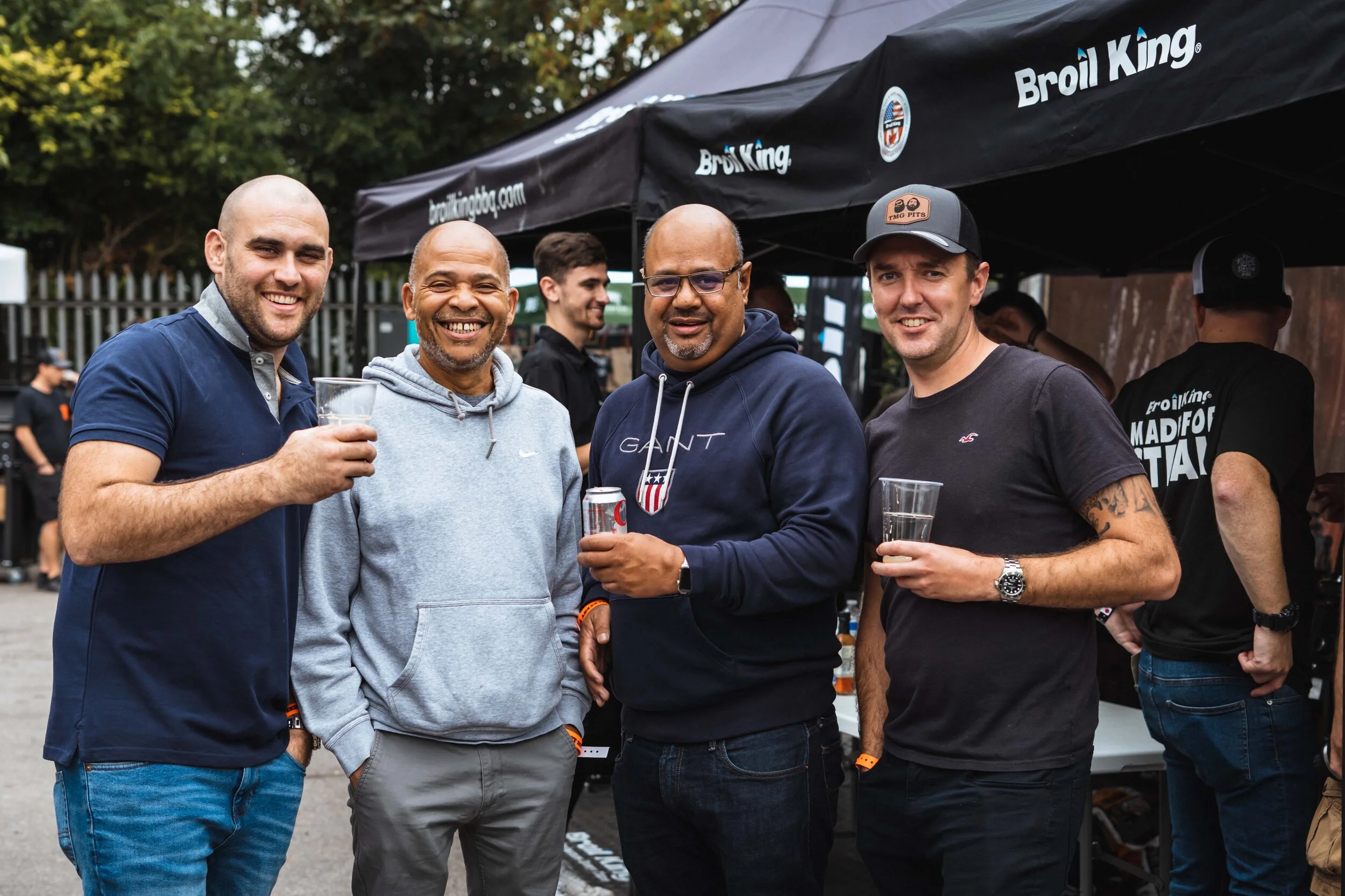 Four smiling men standing outdoors with drinks, under a Broil King tent at an event.