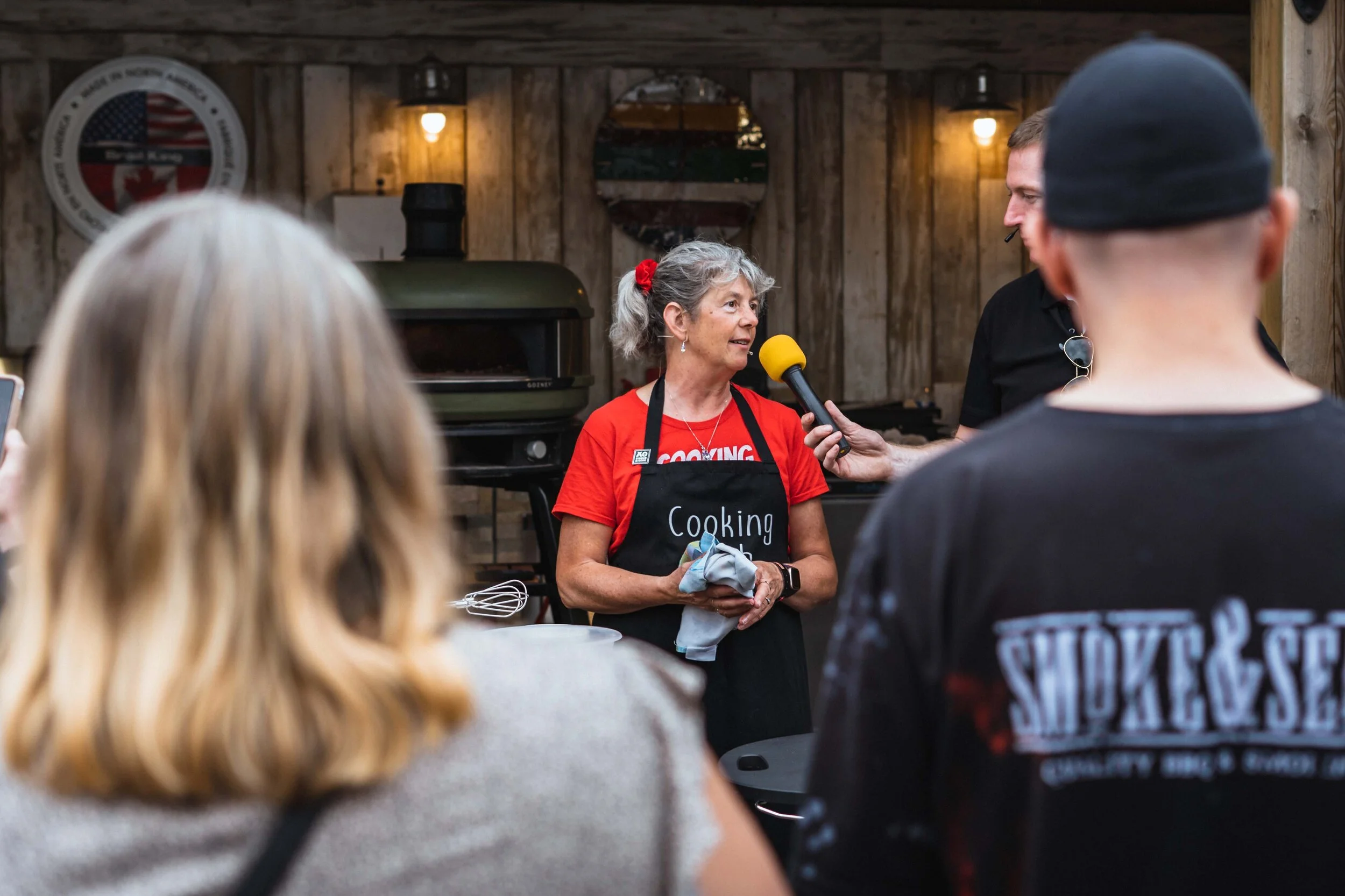 A woman in a red t-shirt and apron is speaking into a microphone held by another person. The setting appears to be an outdoor cooking demonstration. There are people in the foreground watching, and a wood-fired oven in the background.