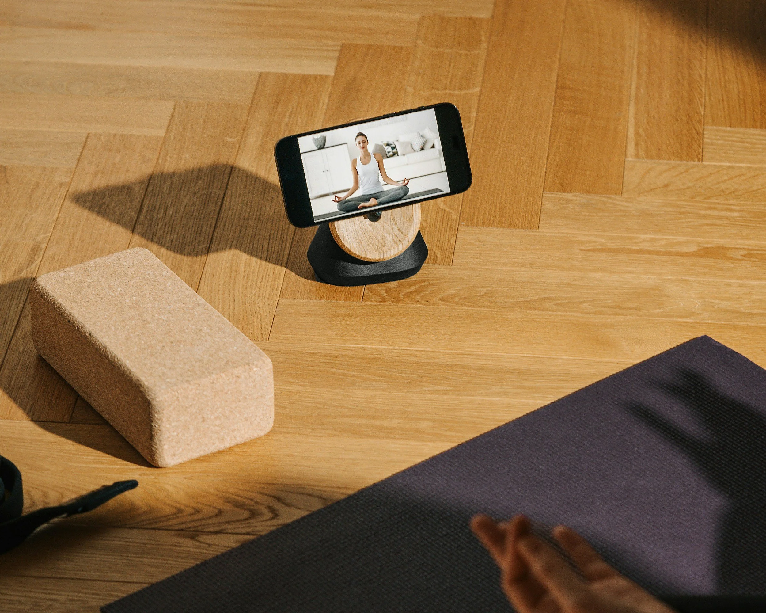 A smartphone on a wooden stand shows a woman meditating in a living room with white furniture and patterned cushions.
