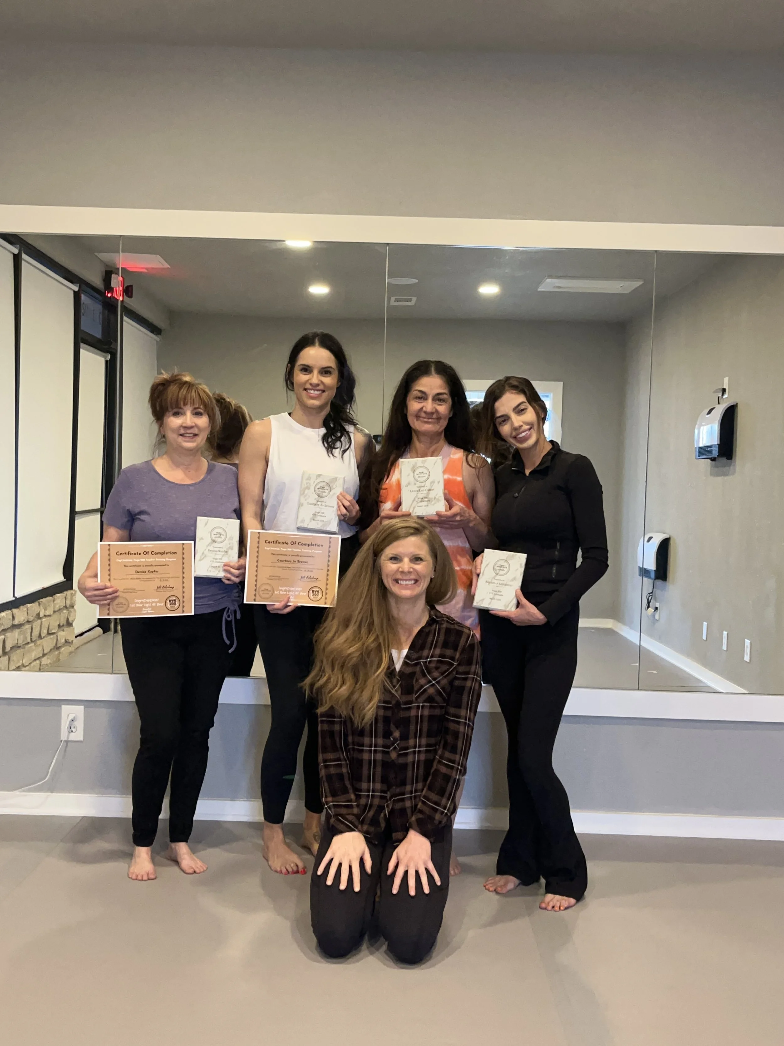 Group of five women posing with certificates and awards in a dance or exercise studio with a large mirror and gray walls.