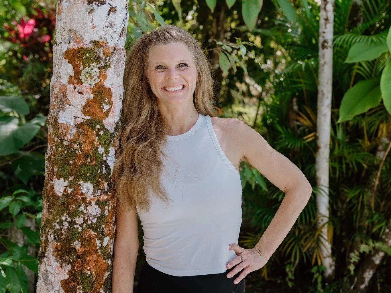 A woman with long wavy hair smiles and stands outdoors next to a tree, surrounded by lush green foliage.