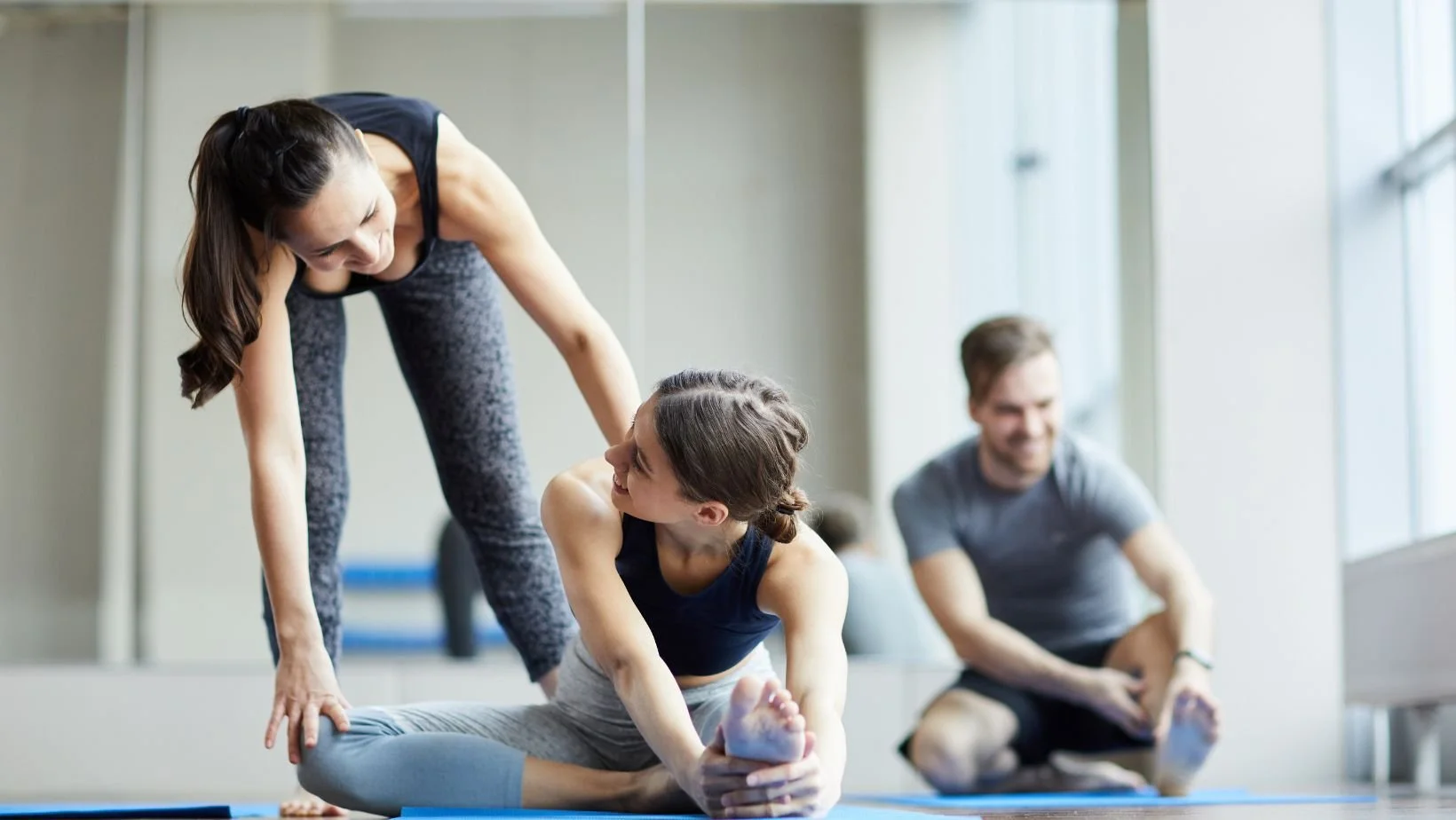 A woman assisting a woman in a yoga pose during a fitness class with a man sitting nearby, in a bright studio with large windows.