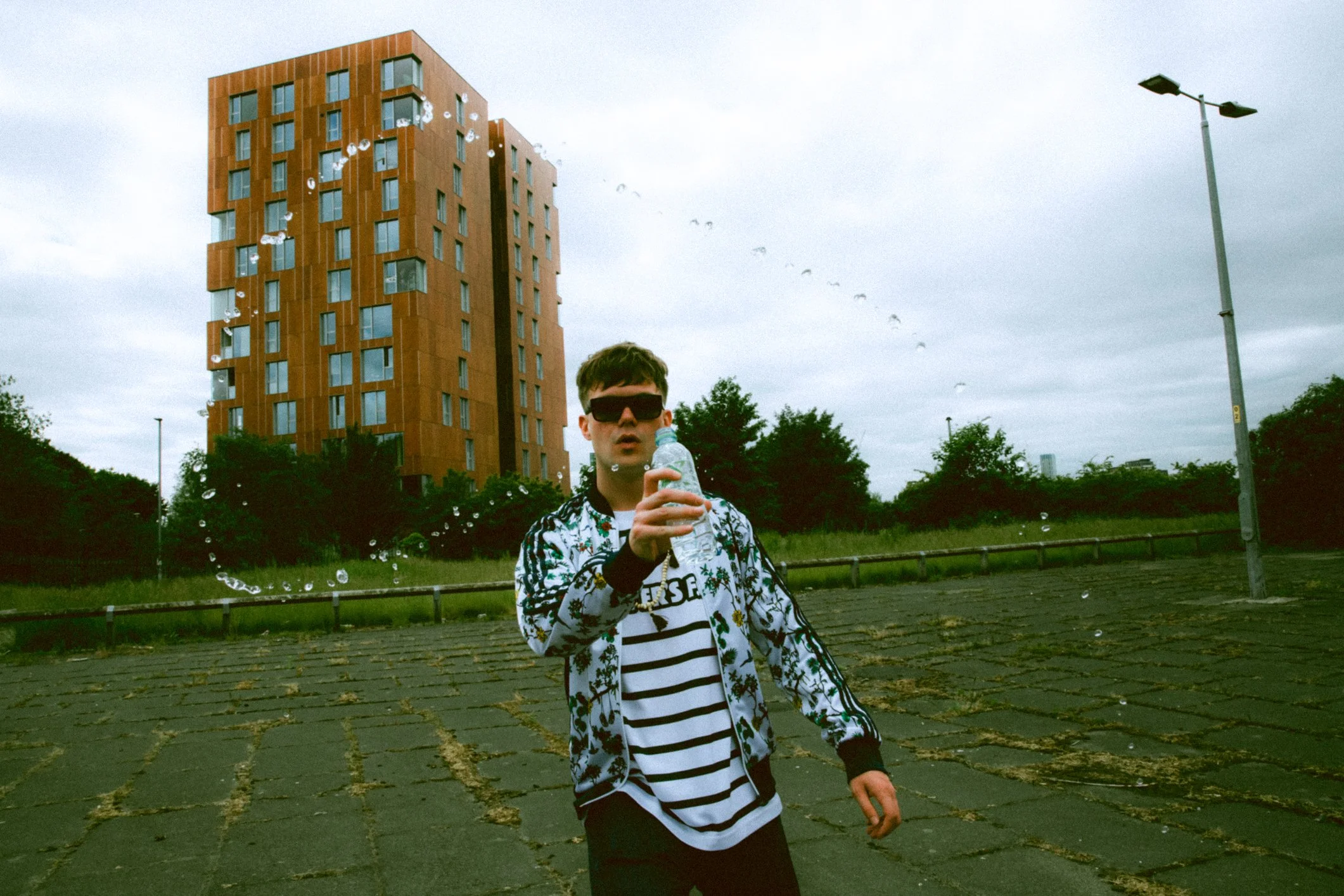 Person wearing sunglasses and a floral jacket, holding a plastic bottle, standing on a paved area with a modern, tall orange building in the background. The scene includes greenery and a cloudy sky.