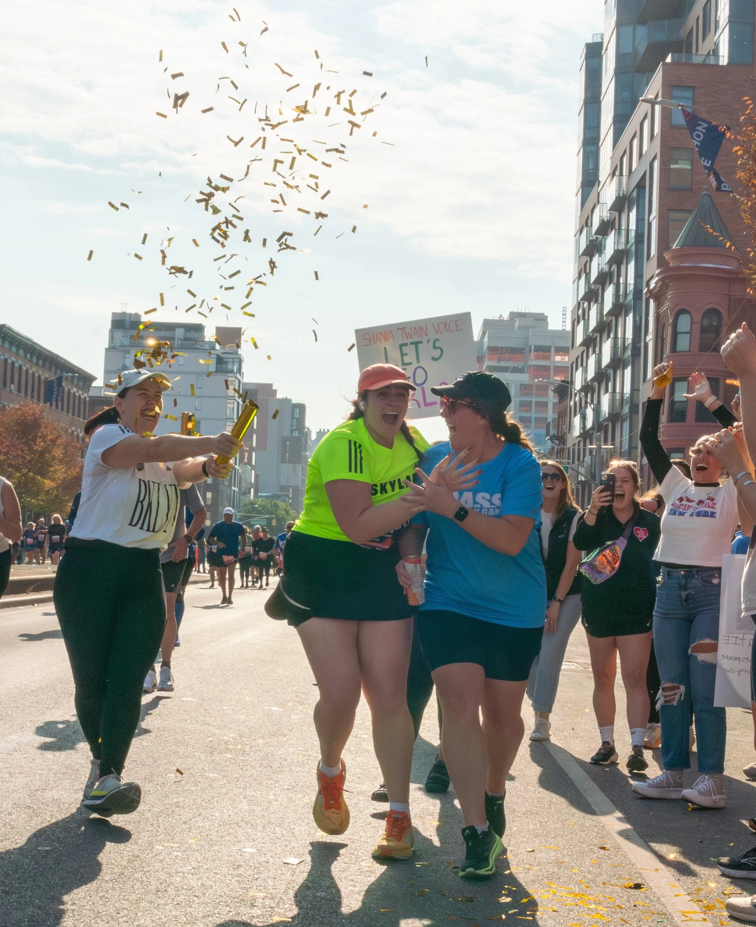 Two BALG runners laugh as a teammate fires a confetti cannon on a city street during a race