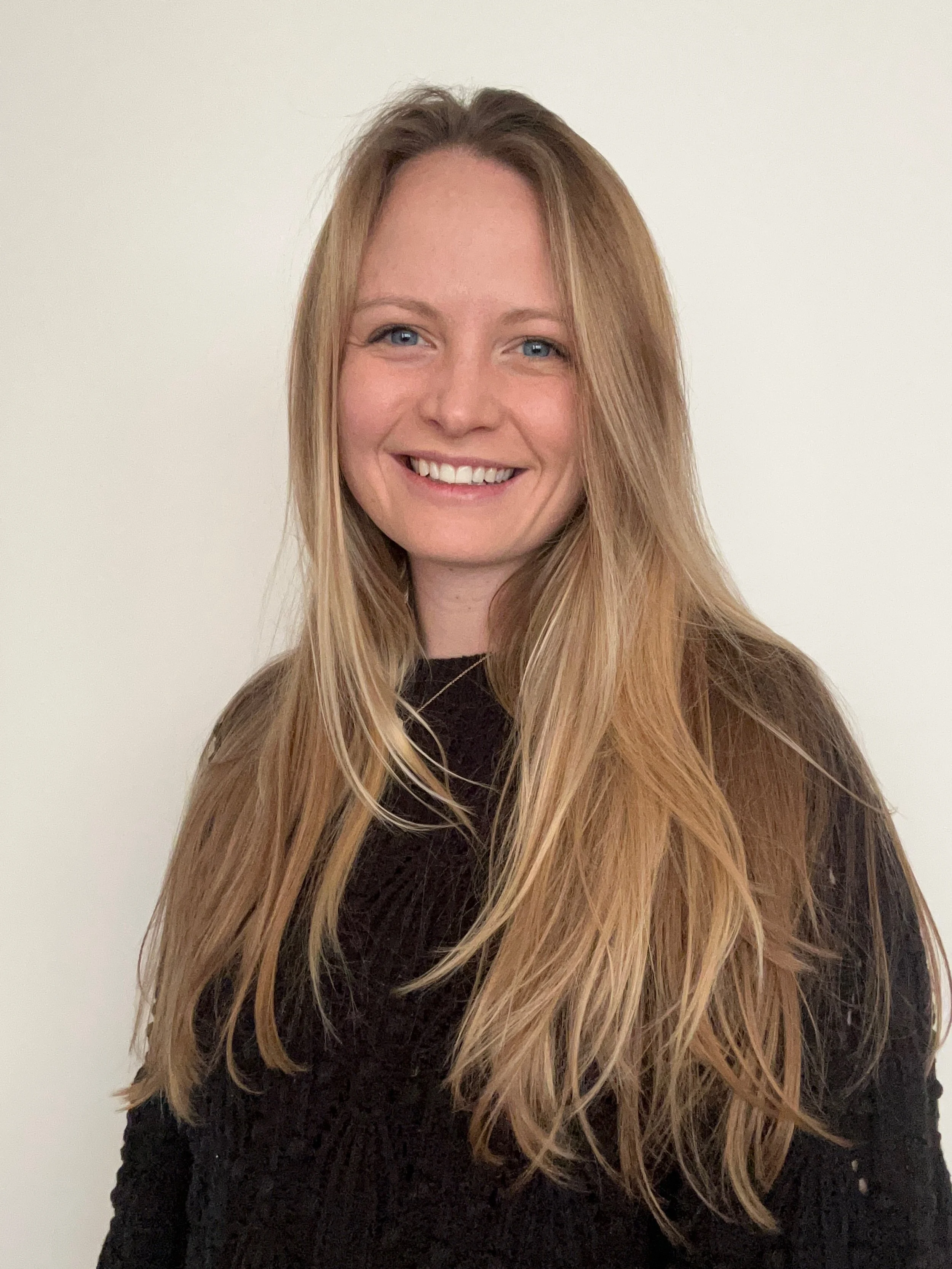 A young woman with long, wavy blonde hair, blue eyes, and a wide smile, wearing a black sweater, standing against a plain white background.