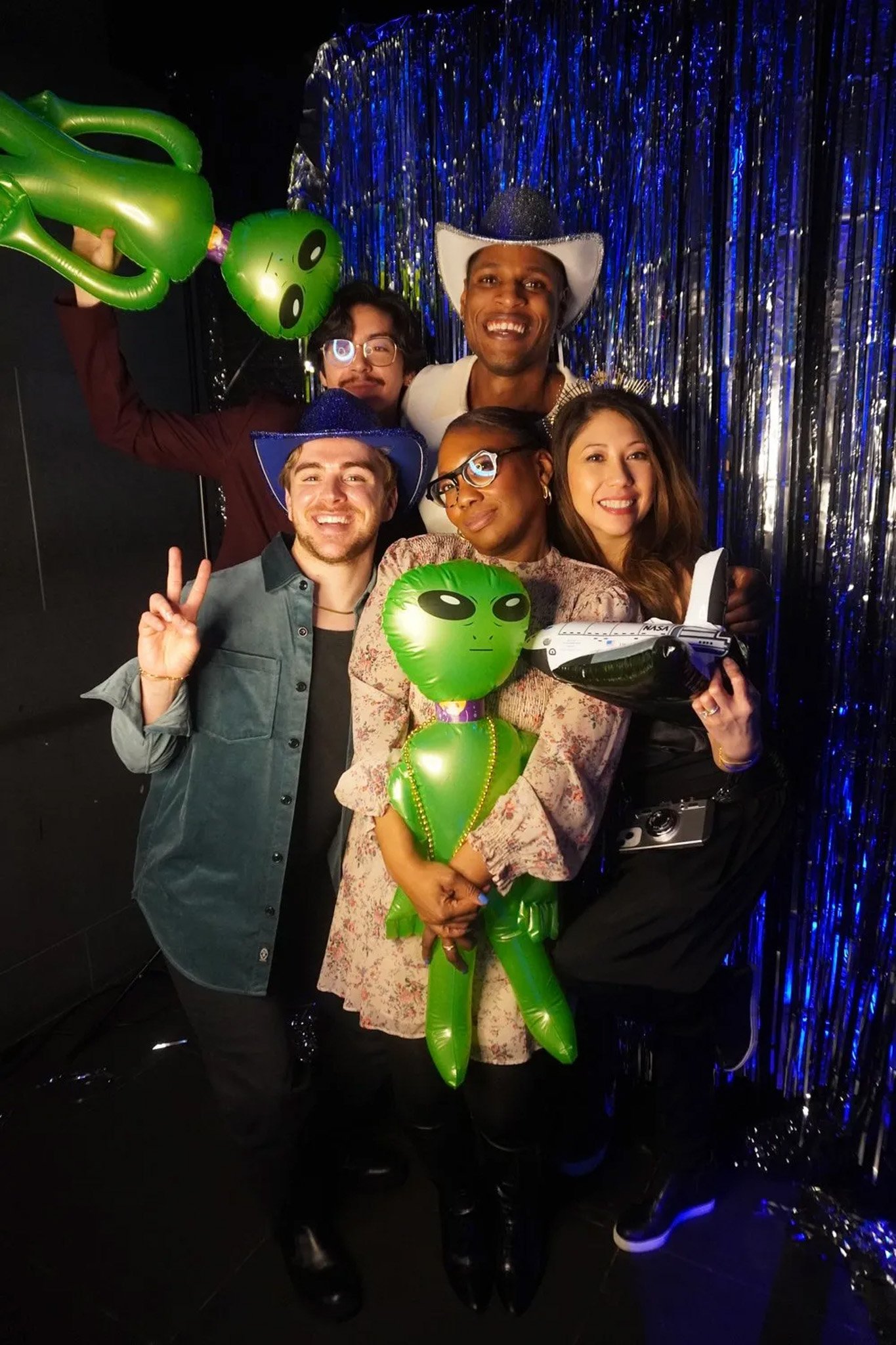 Group of people at a space-themed party posing with inflatable aliens, wearing cowboy hats and holding a toy space shuttle. They are standing in front of a metallic fringe curtain.