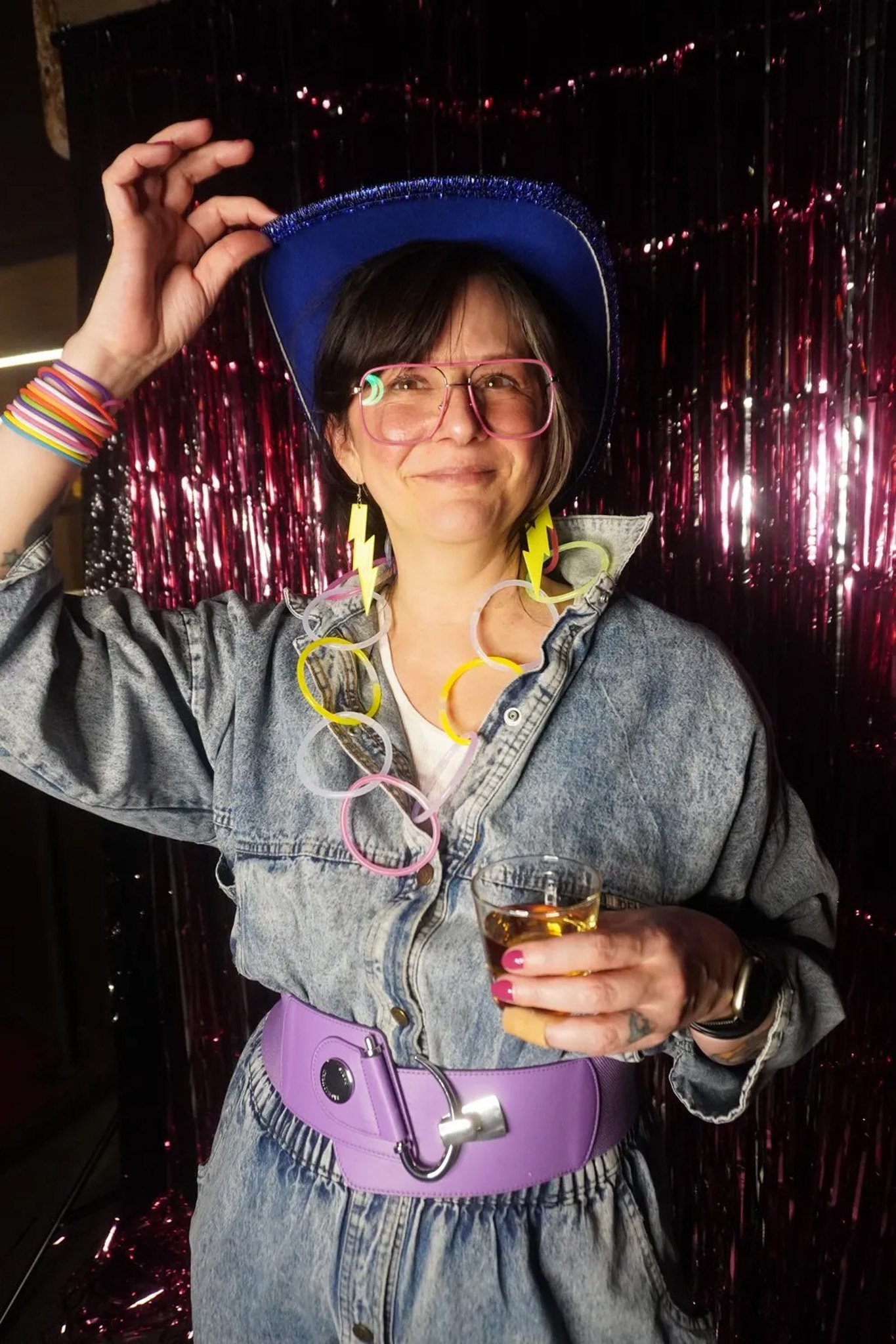 Person in denim outfit with a blue hat and colorful accessories, including neon bracelets and earrings, holding a drink in front of a shiny backdrop.