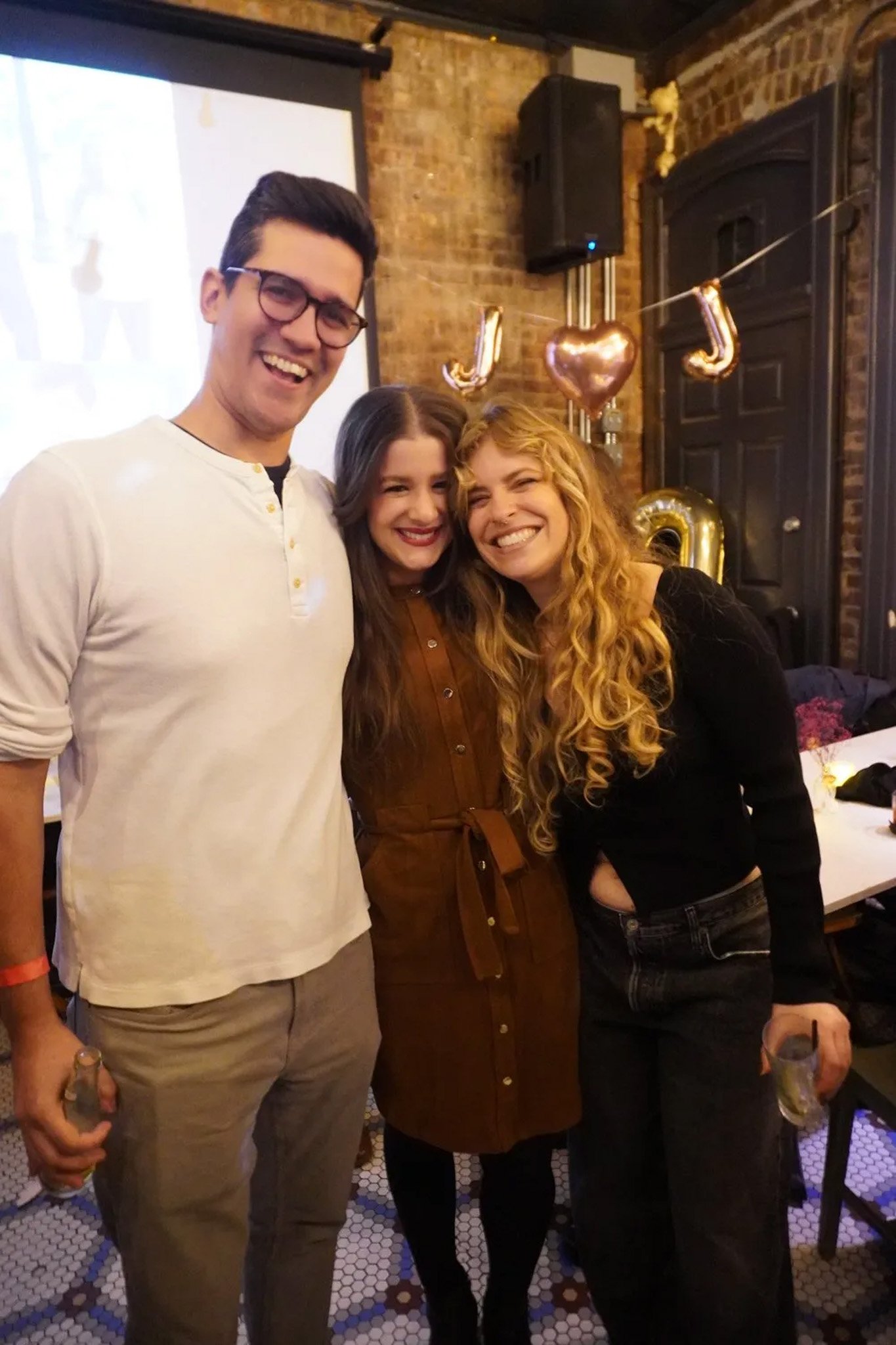 Three people smiling at a party with balloons forming a heart and letters J and J.