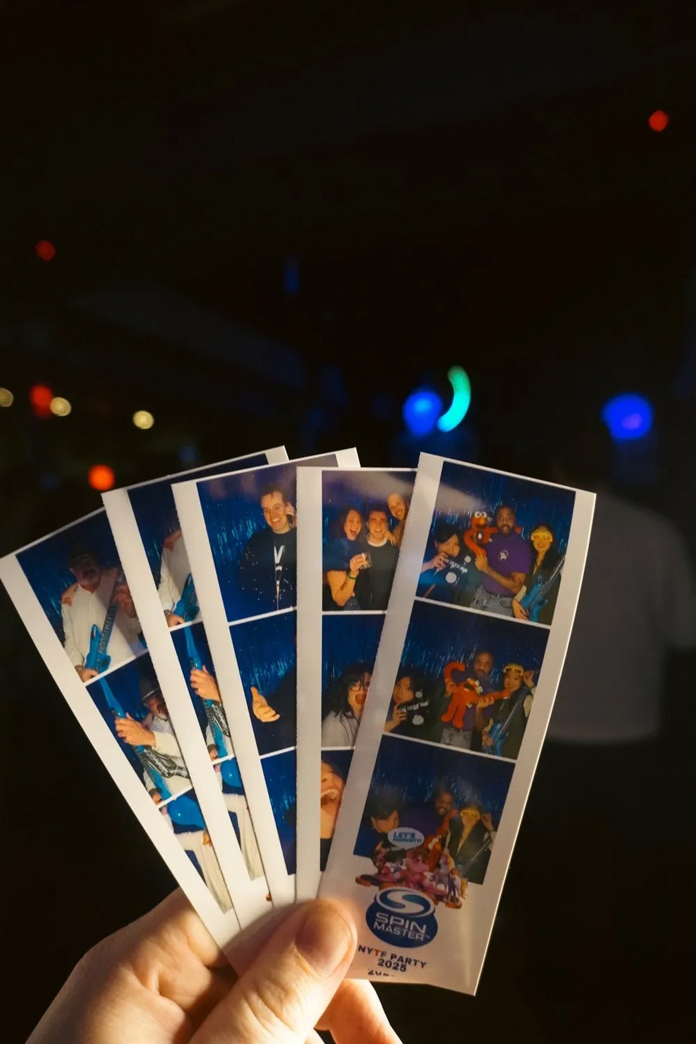 Person holding photo strips from a party with people posing, some with toys and props, a logo and "Spin Master NYTF Party 2023" printed at the bottom.