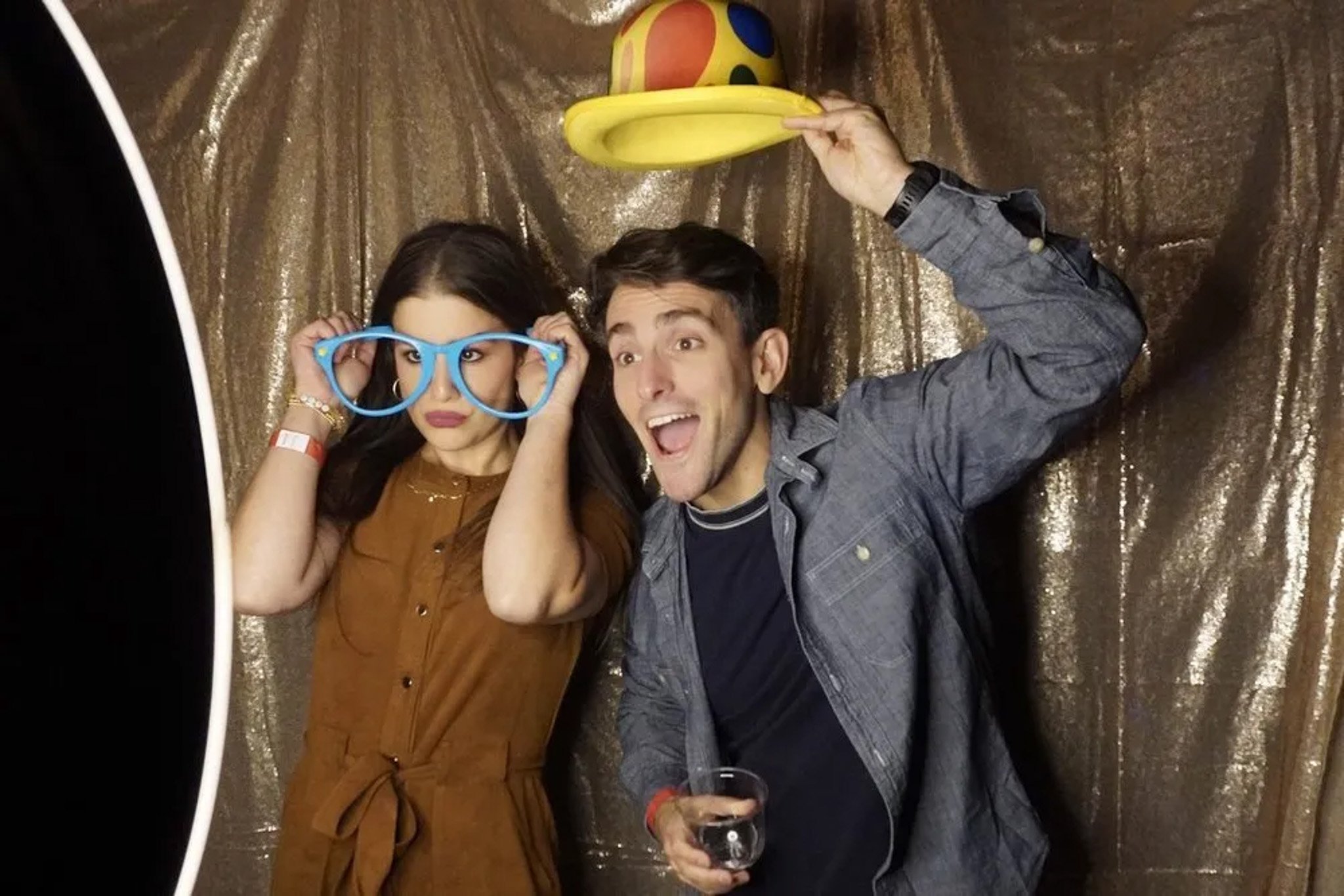 Two people posing in a photo booth. The woman wears oversized blue glasses, and the man holds a colorful clown hat above his head, smiling. A golden backdrop is behind them.