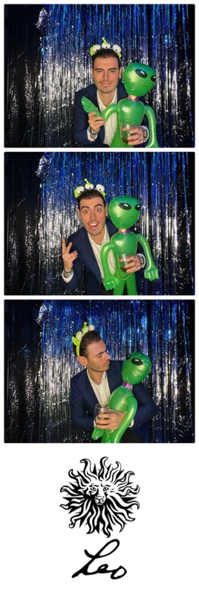 Man posing with inflatable alien and wearing alien-themed headband, in front of a shiny curtain backdrop, holding a drink, with a stylized Leo sign and lion face at the bottom.