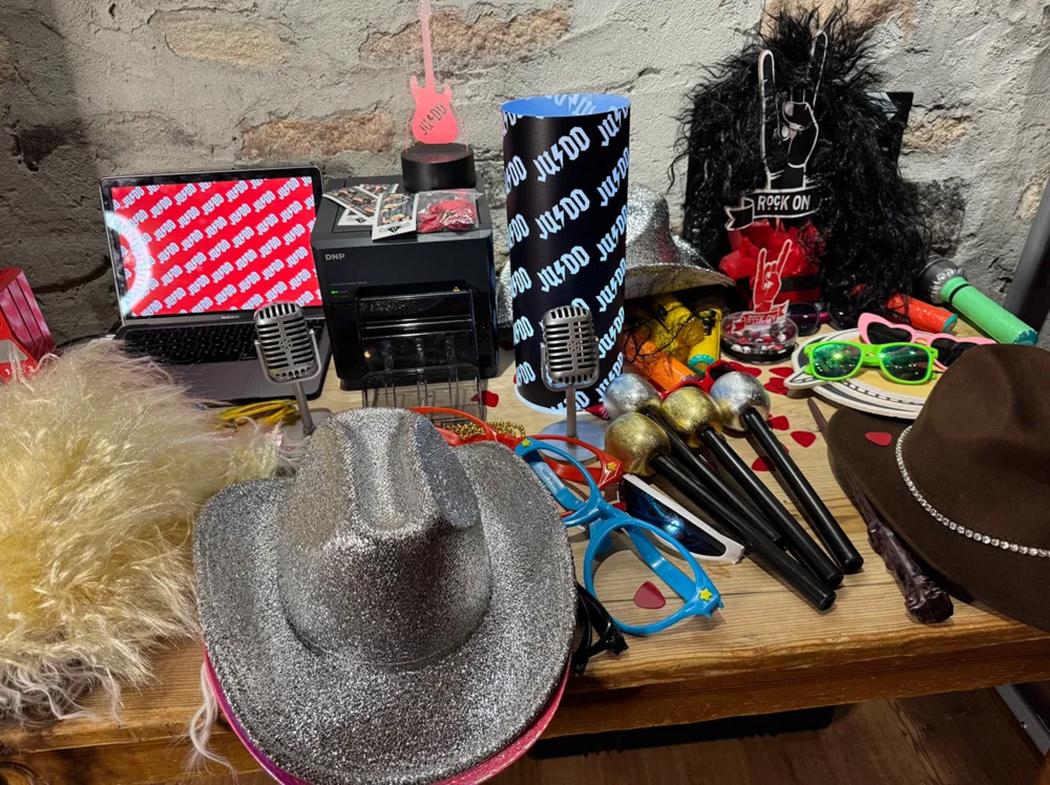 Variety of party props on a table, including glitter hats, novelty sunglasses, microphones, and a laptop in front of a brick wall.