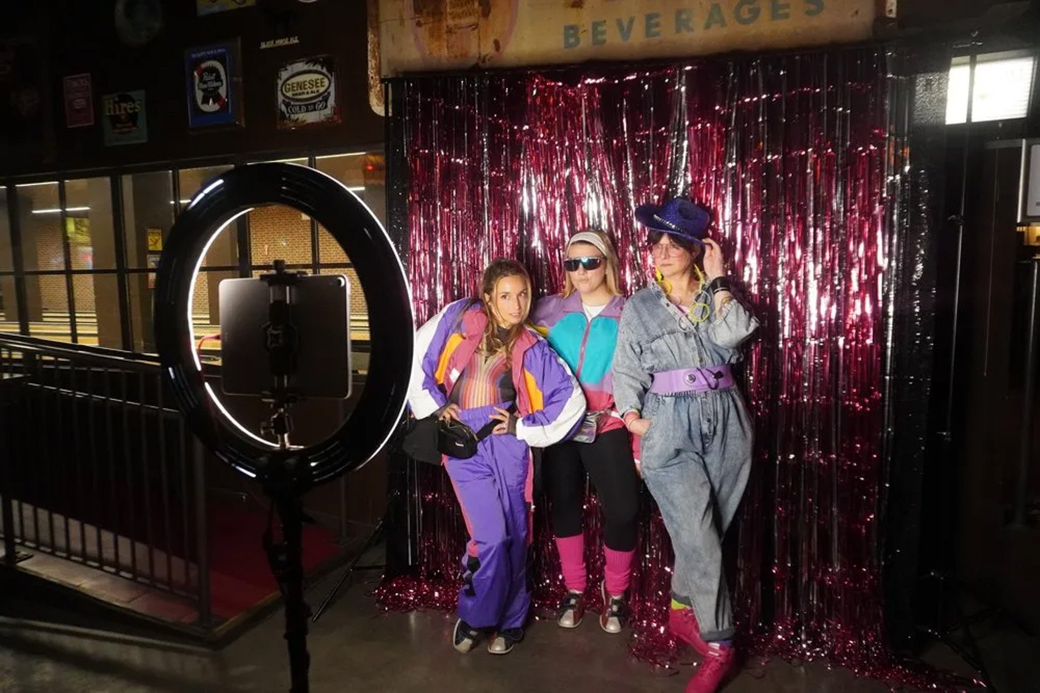 Three people wearing retro 80s outfits pose in front of a shiny pink backdrop, with a ring light setup for photography in the foreground.