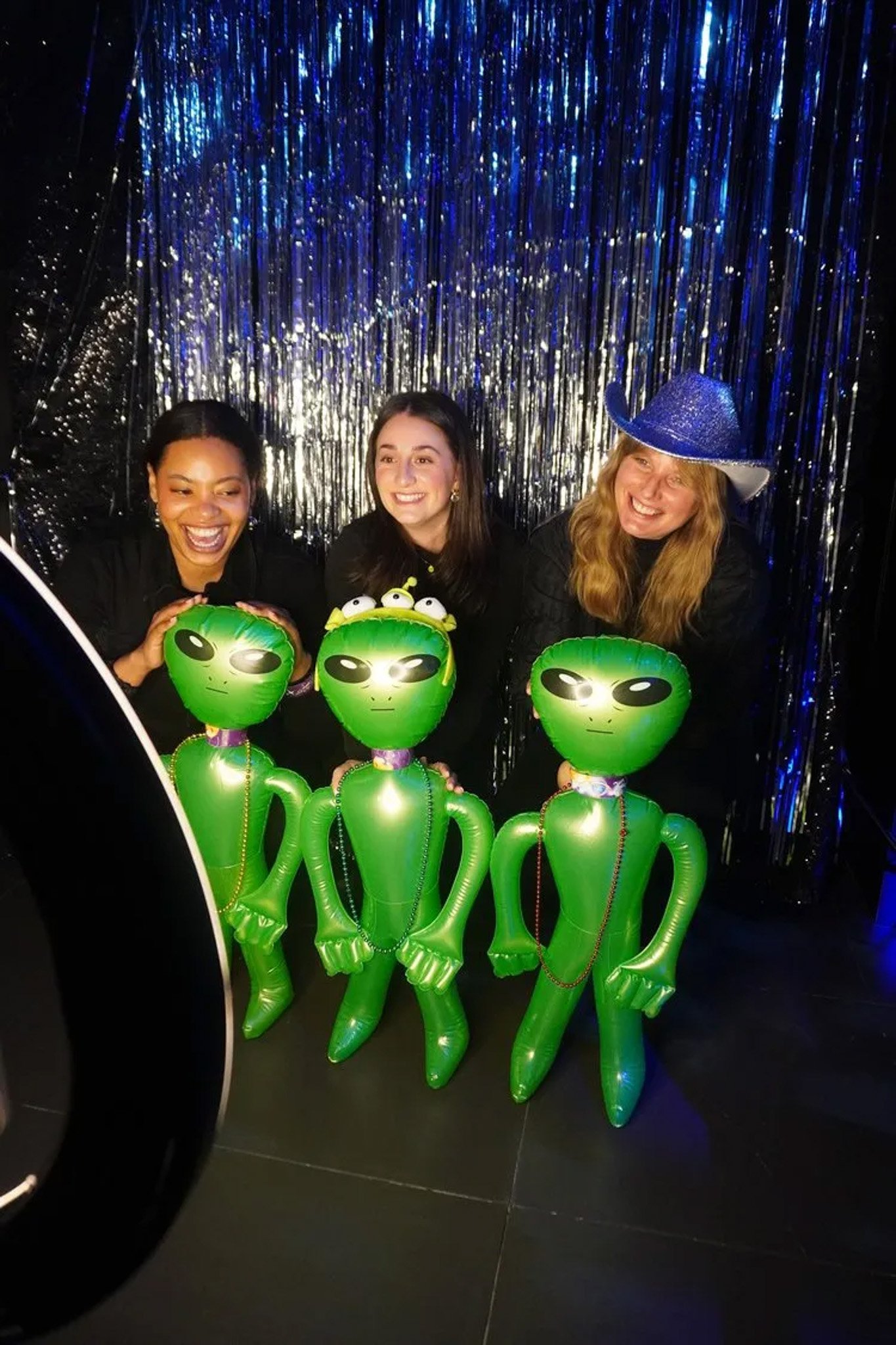 Three people posing with inflatable alien toys in front of a sparkly backdrop. One person is wearing a blue cowboy hat.