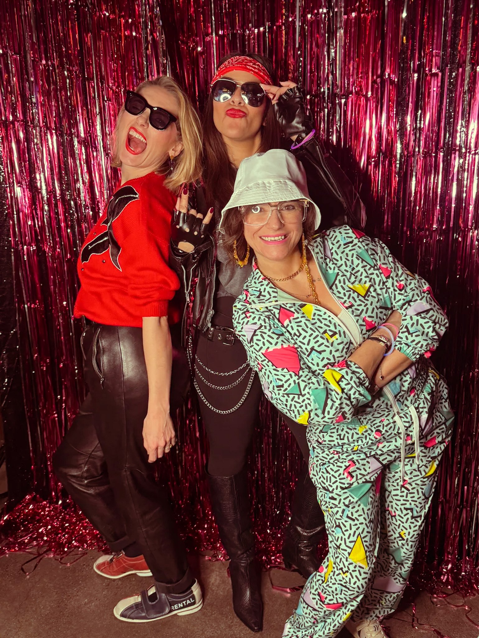 Three women posing in front of a shiny pink backdrop, wearing retro-inspired outfits including sunglasses, a red sweater, leather pants, a bandana, a bucket hat, and a colorful tracksuit.
