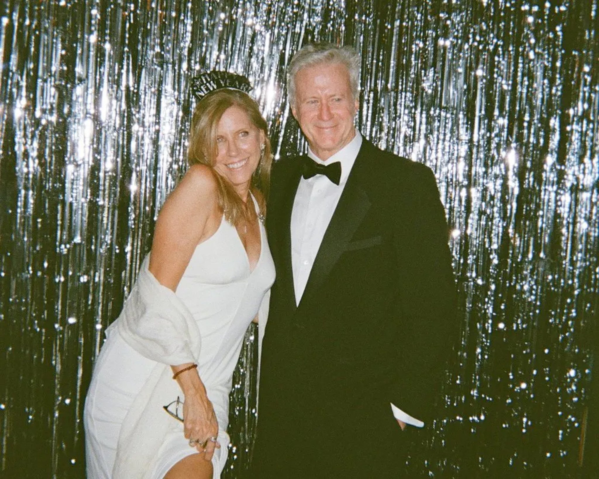 A woman in a white dress and a man in a tuxedo posing in front of a shimmering curtain backdrop, celebrating New Year's Eve.