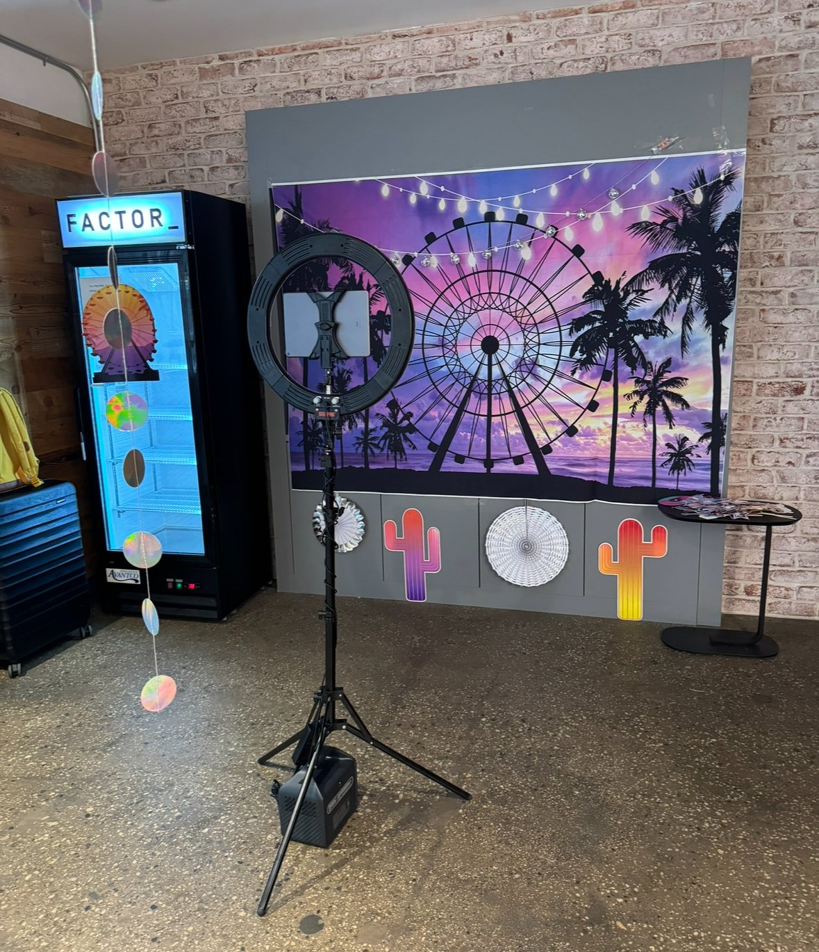 Photobooth setup with a colorful Ferris wheel backdrop, ring light on tripod, decorative fridge with "FACTOR" sign, brick wall, and cactus and paper wheel decorations.