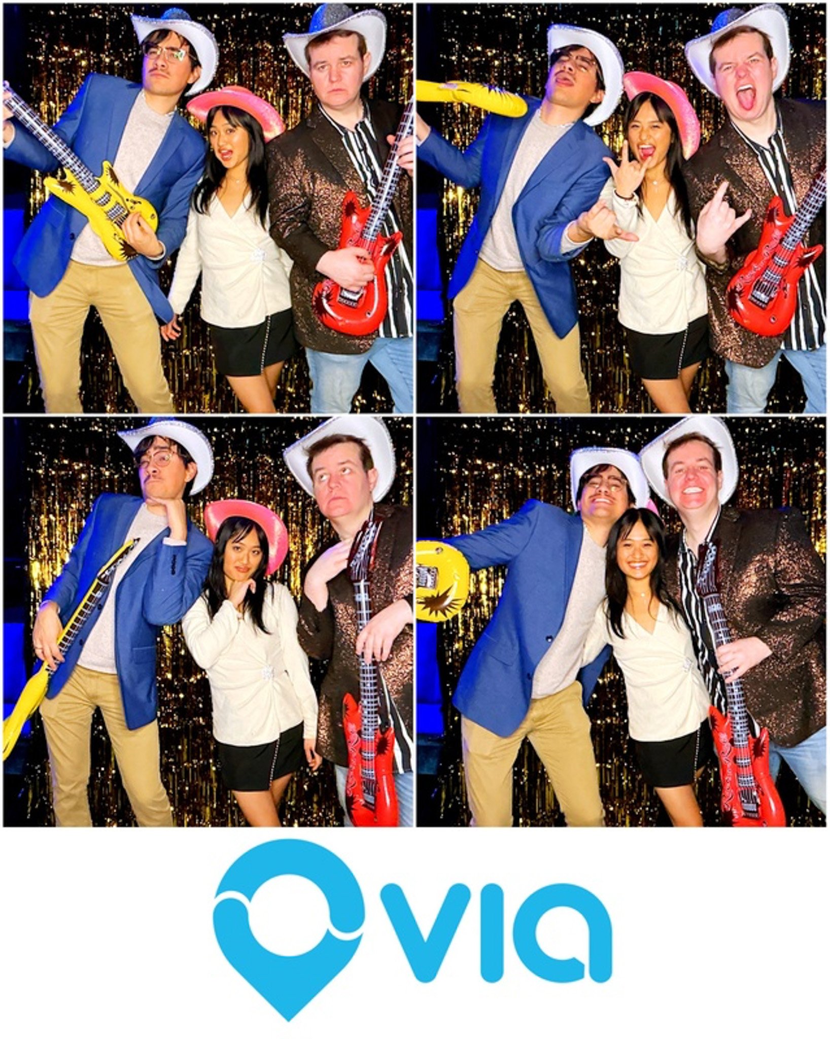 Three people wearing cowboy hats posing in a photo booth with inflatable guitars, smiling and making fun gestures, in front of a glittery backdrop. The Via logo is displayed at the bottom.