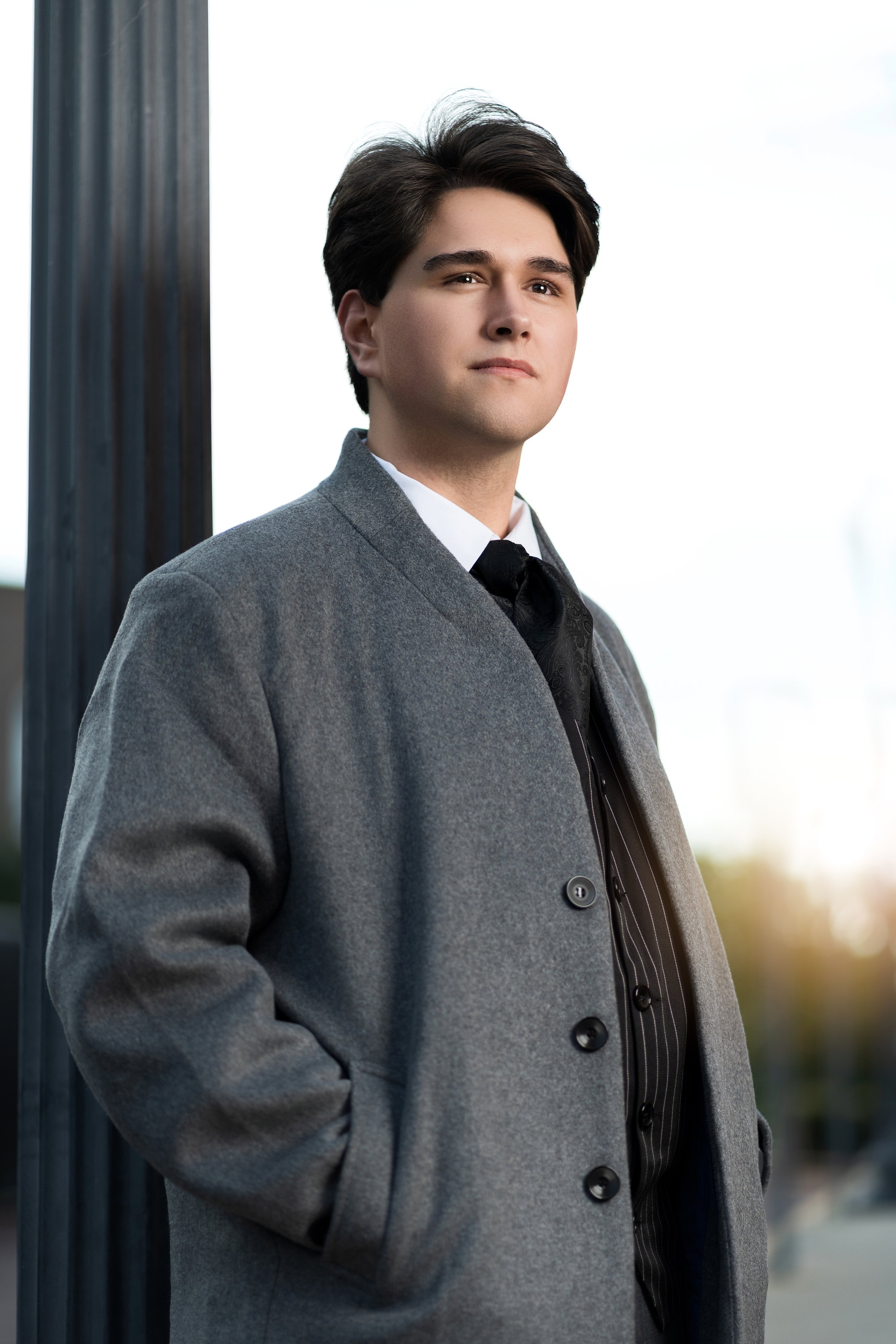 A young man wearing a gray coat over a suit with a white shirt and black tie, standing outdoors near a black pole, with a blurred background and sunlight coming from the right.