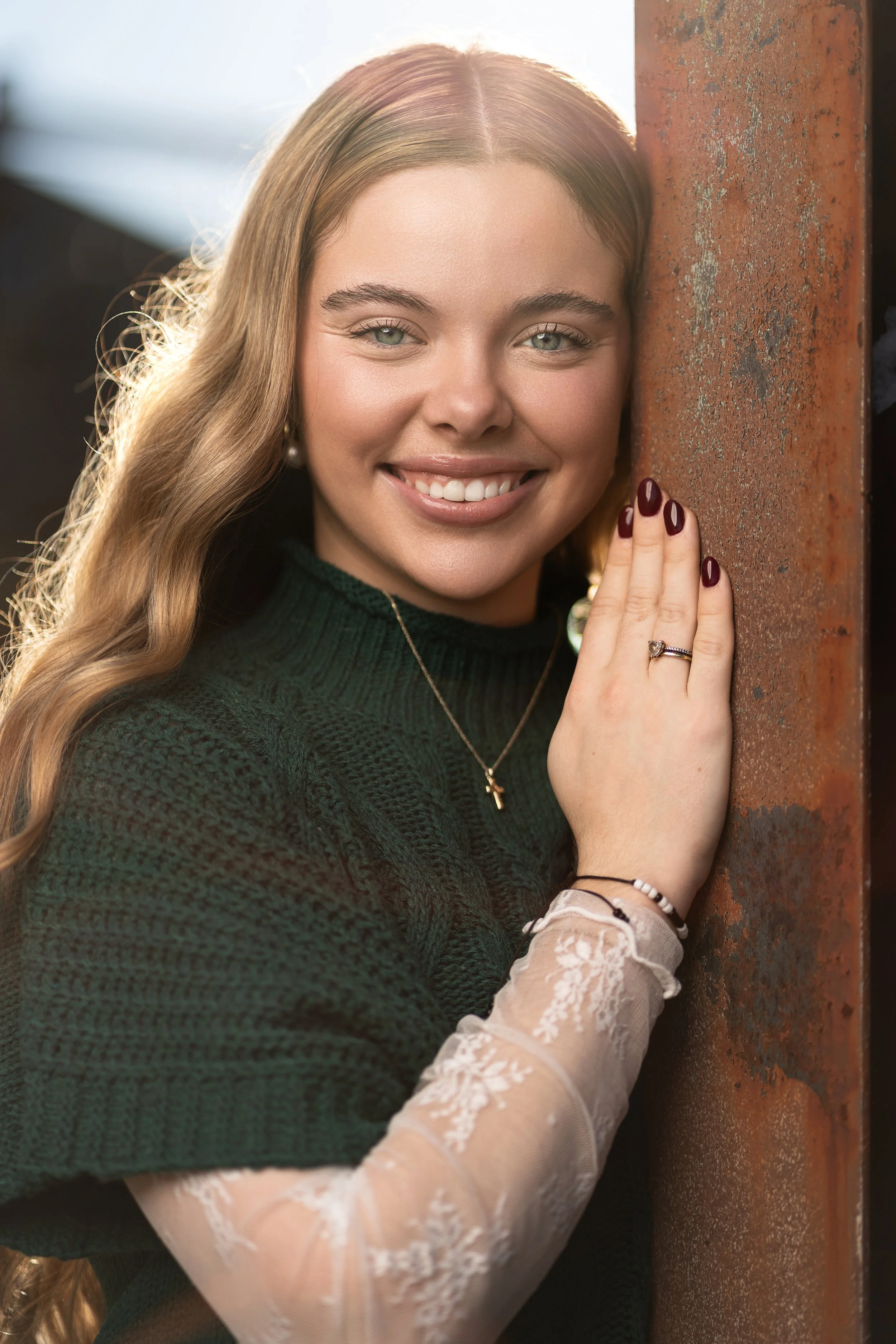 A young woman with long, wavy blonde hair smiles as she leans against a rust-colored metal surface, wearing a dark green sweater and lace-embellished sheer sleeve, accessorized with necklaces, rings, and bracelets.