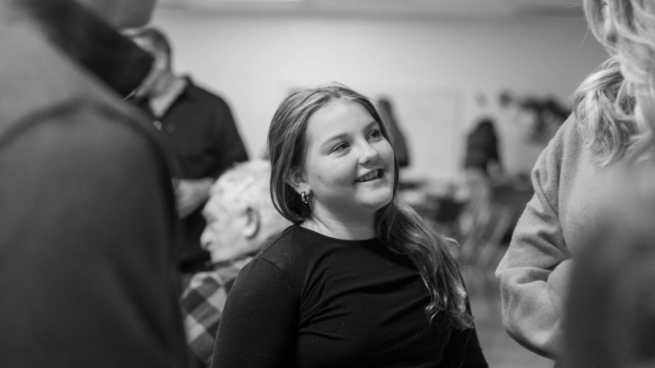 A young woman with long hair smiling and listening in a conversation in a social setting, with other people in the background.
