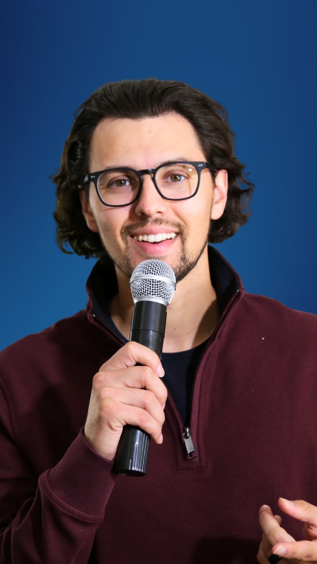 A man with glasses holding a microphone, smiling and speaking.
