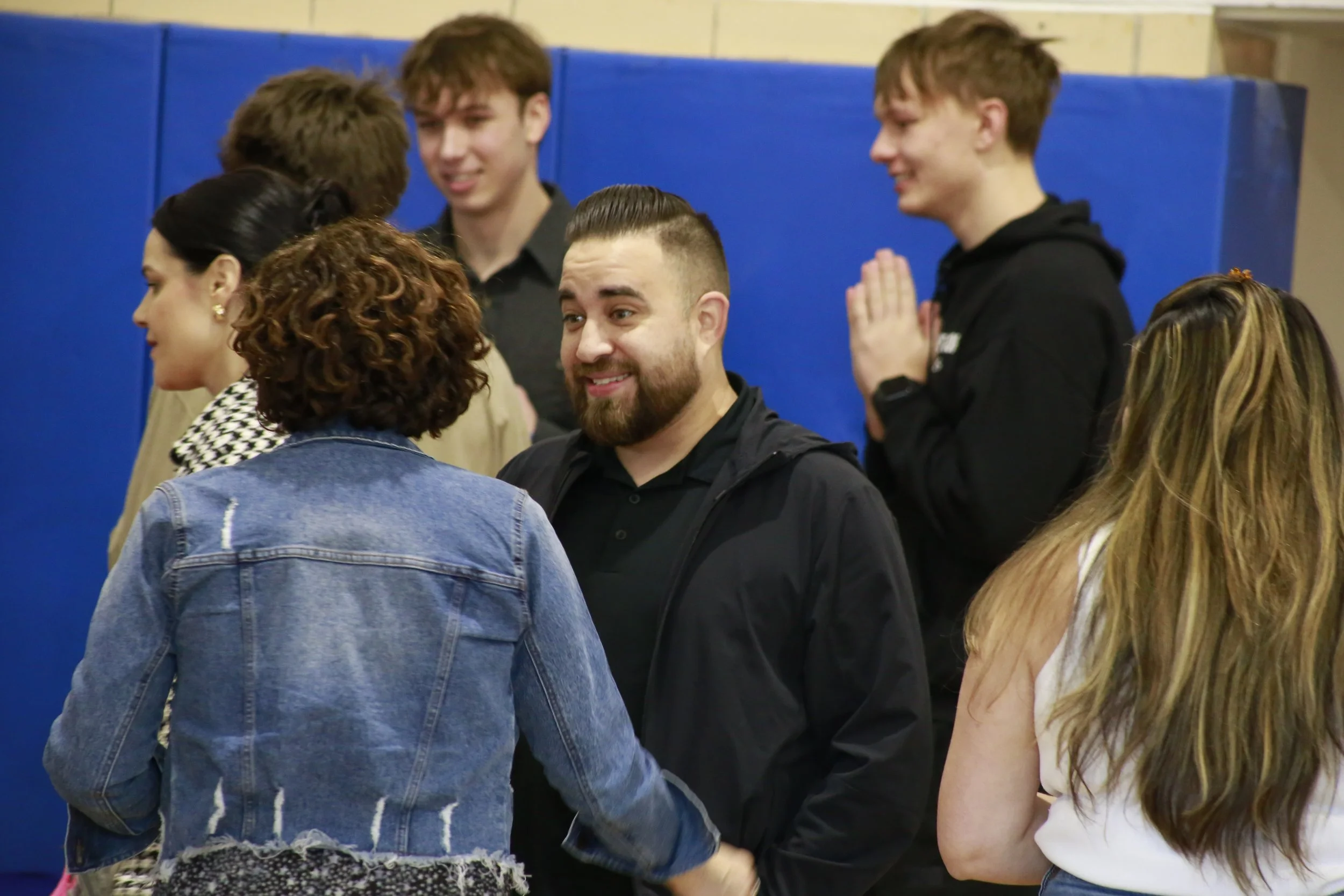 A man with glasses, a bald head, and a white beard, wearing a checkered shirt, is talking to a person with curly hair, wearing a black t-shirt with white text, in front of blue curtains.