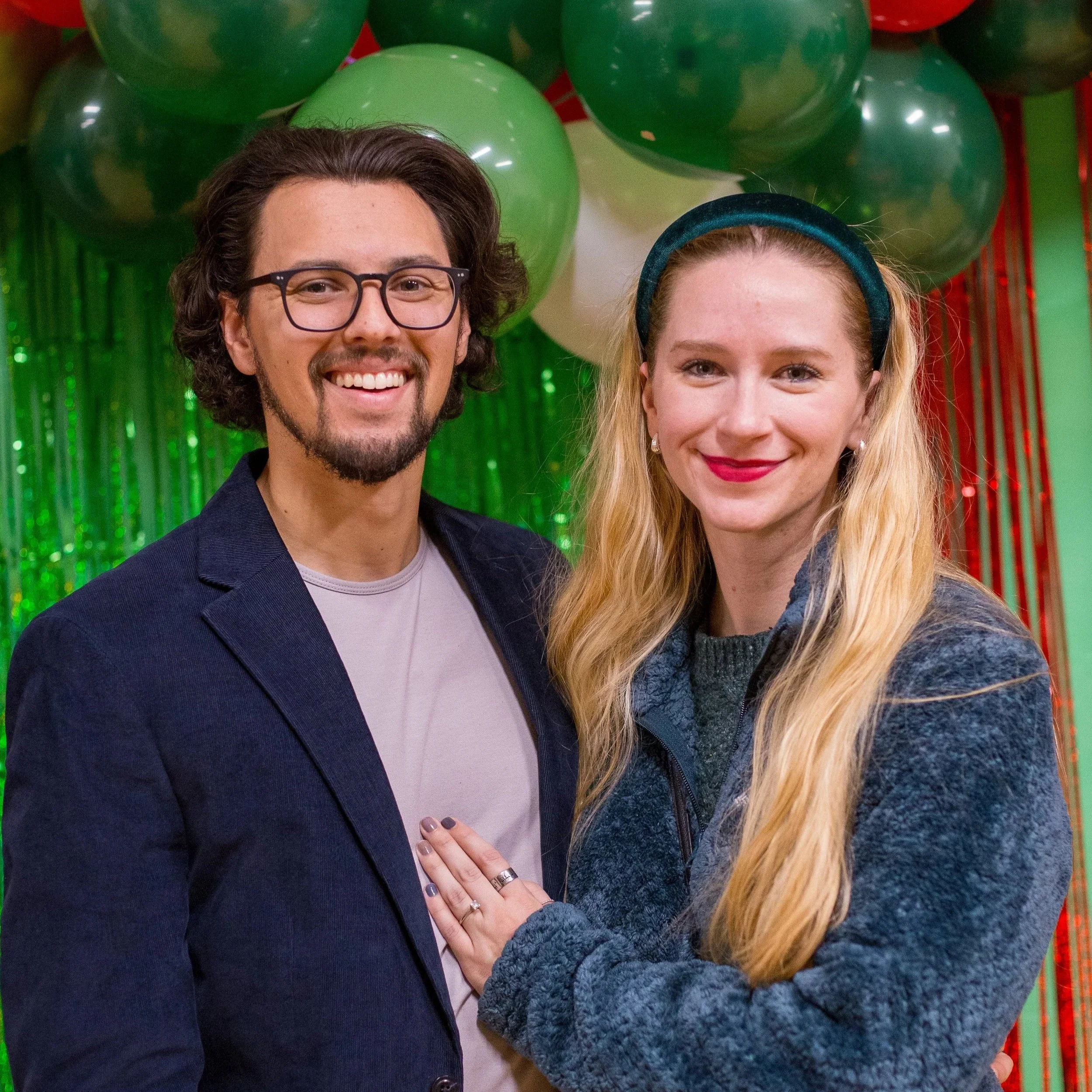 A smiling man and woman standing close together at party with green and red balloons and festive decorations in the background.