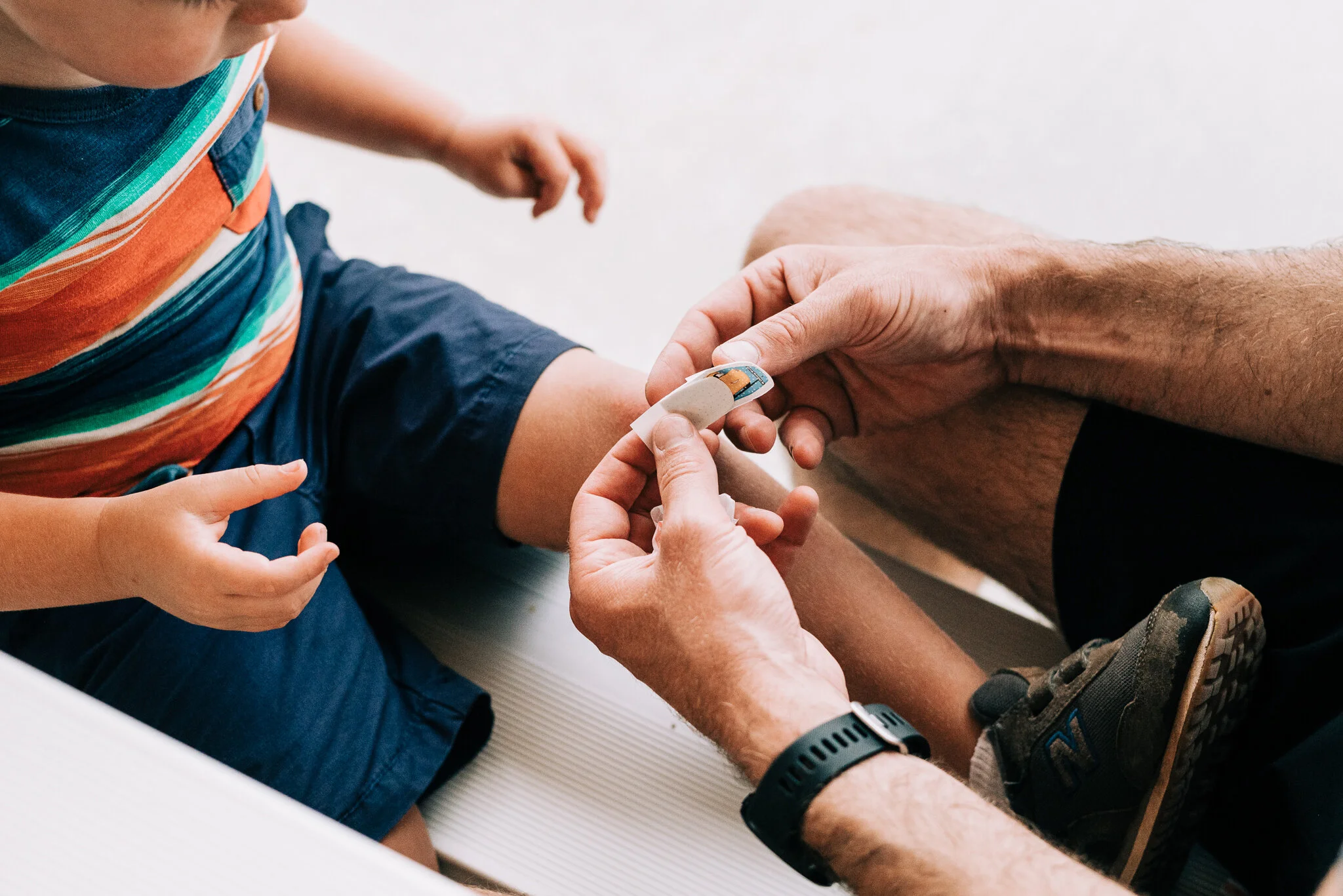 A young child receiving a bandage on his knee from an adult, with another child's hand reaching out nearby.