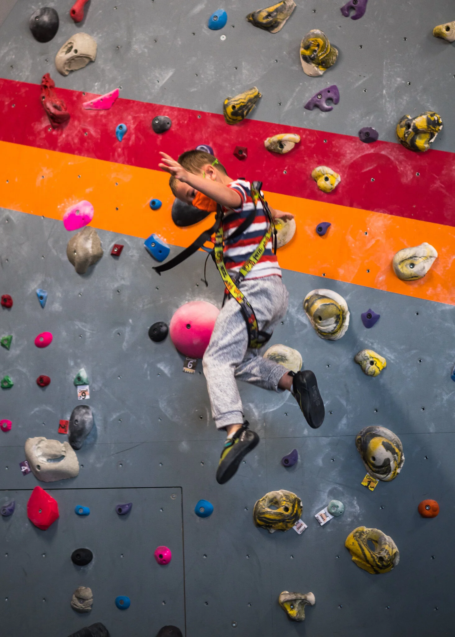 A young boy climbing an indoor rock wall with colorful holds and a harness.