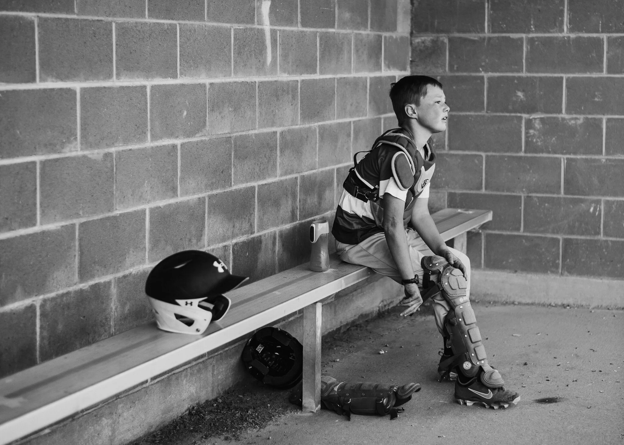 A young boy sitting on a bench in a baseball uniform with protective gear, including a helmet and cleats, looking contemplative. Sports equipment and water bottles are next to him. The background features a brick wall.