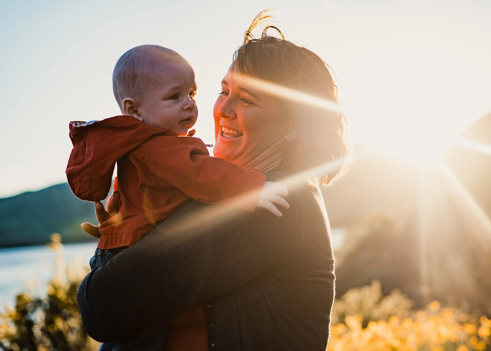 A woman smiling and holding a young child outdoors during sunset.