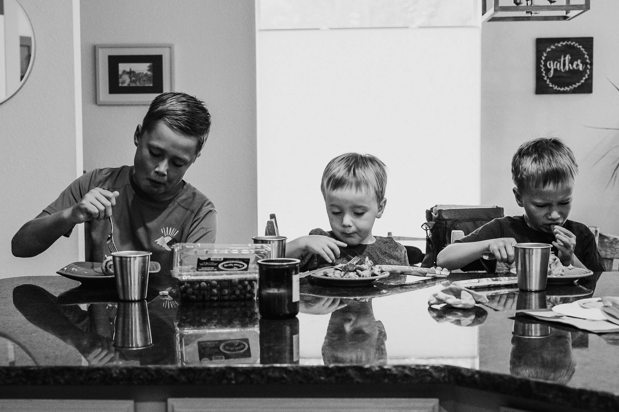 Three young boys sitting at a dining table, eating a meal together, with food, cups, and snacks on the table, in a home setting.
