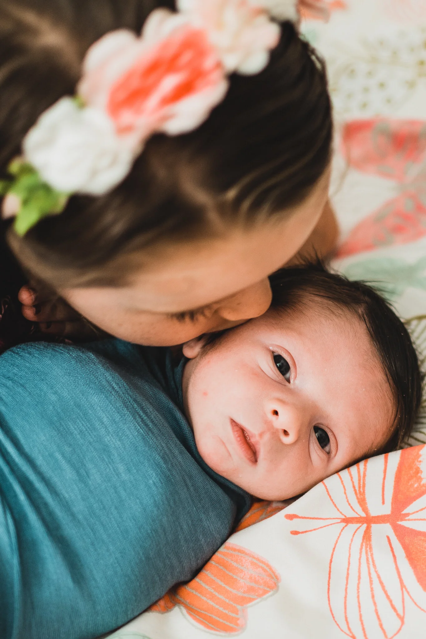A woman is lying down and kissing a sleeping baby on the forehead. The baby is lying on a pillow with a butterfly pattern.