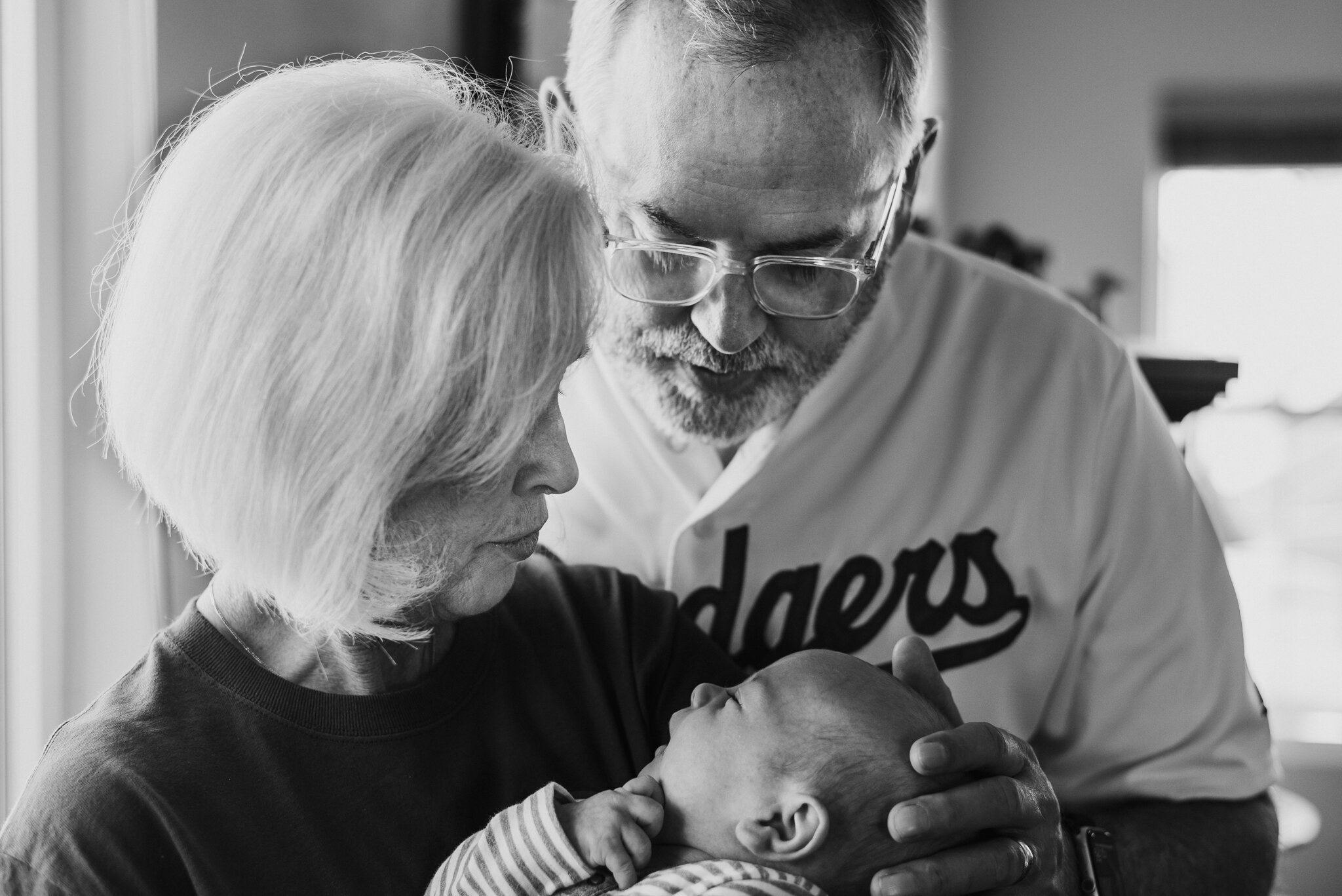 An elderly woman and a man with glasses, likely her son, are holding a newborn baby. The woman and the man are looking lovingly at the baby, who is lying down, gazing back. The scene is intimate and tender.