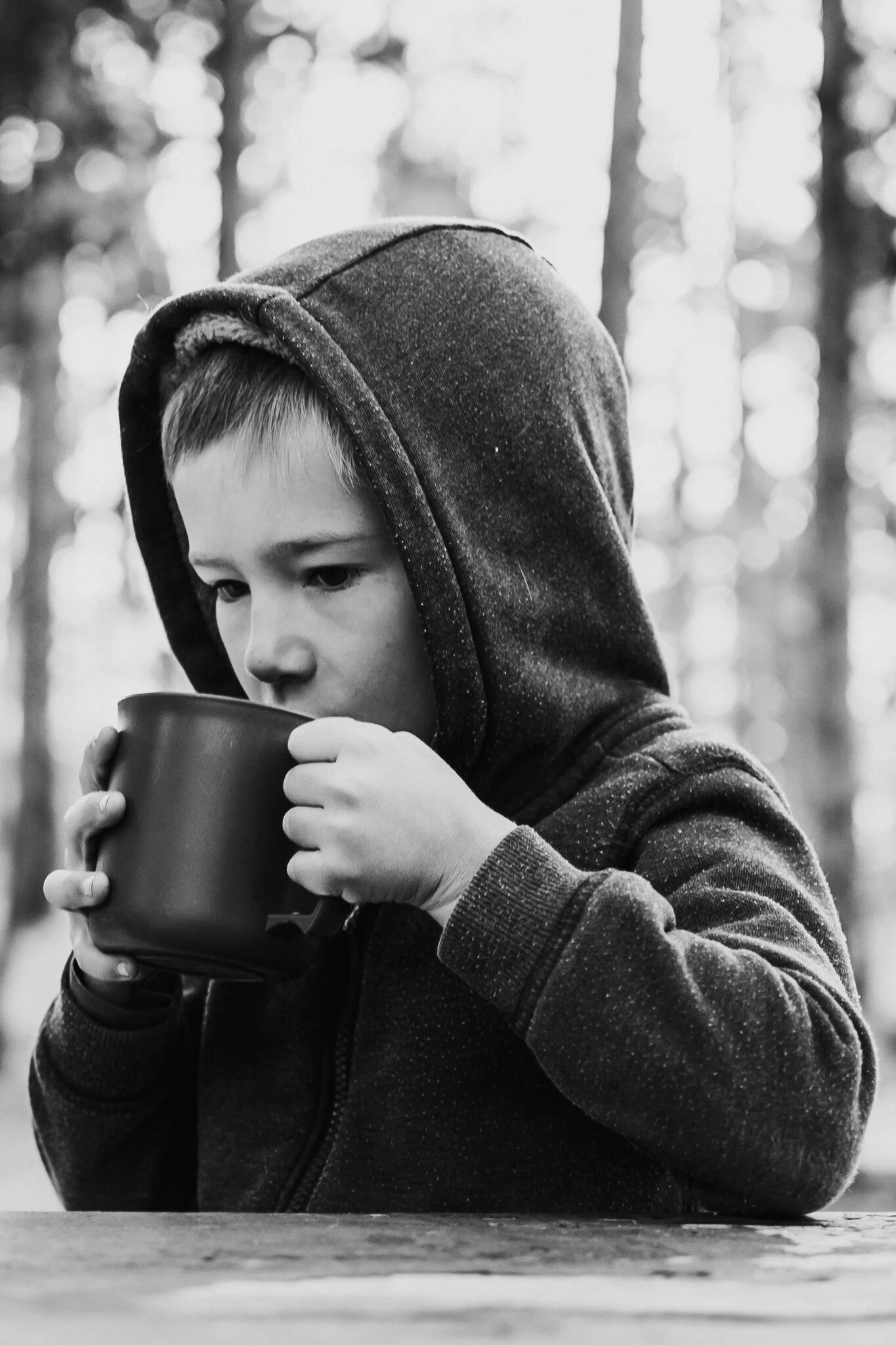A young boy in a hoodie drinking from a mug outdoors.