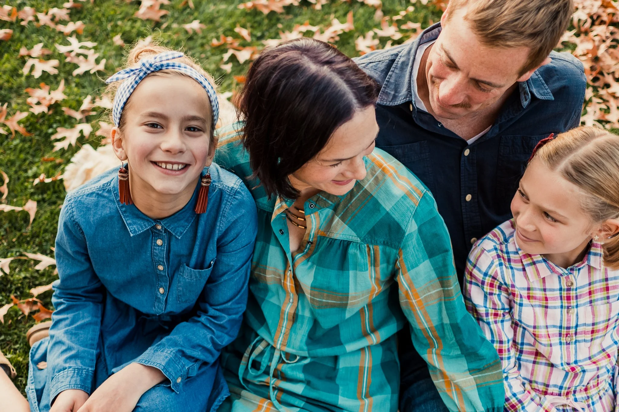 A family of four sitting outdoors on grass with fallen leaves, smiling and looking at each other.