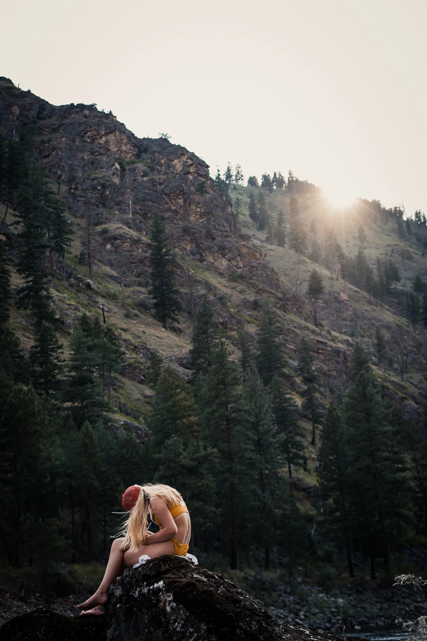 A girl with long blonde hair wearing a yellow dress and a red hat sitting on a rock in a forested mountainous area during sunset.