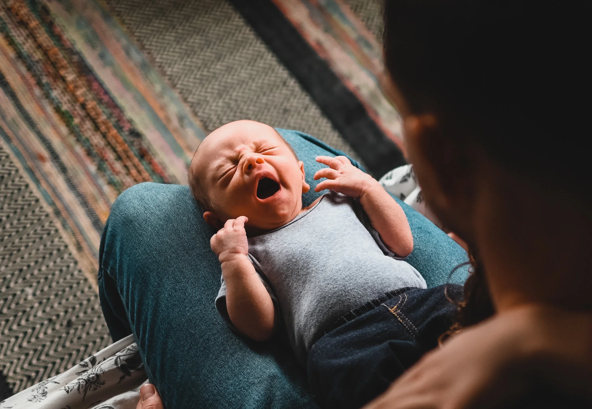 A baby crying while lying on an adult's lap, with the adult sitting on a patterned carpet.