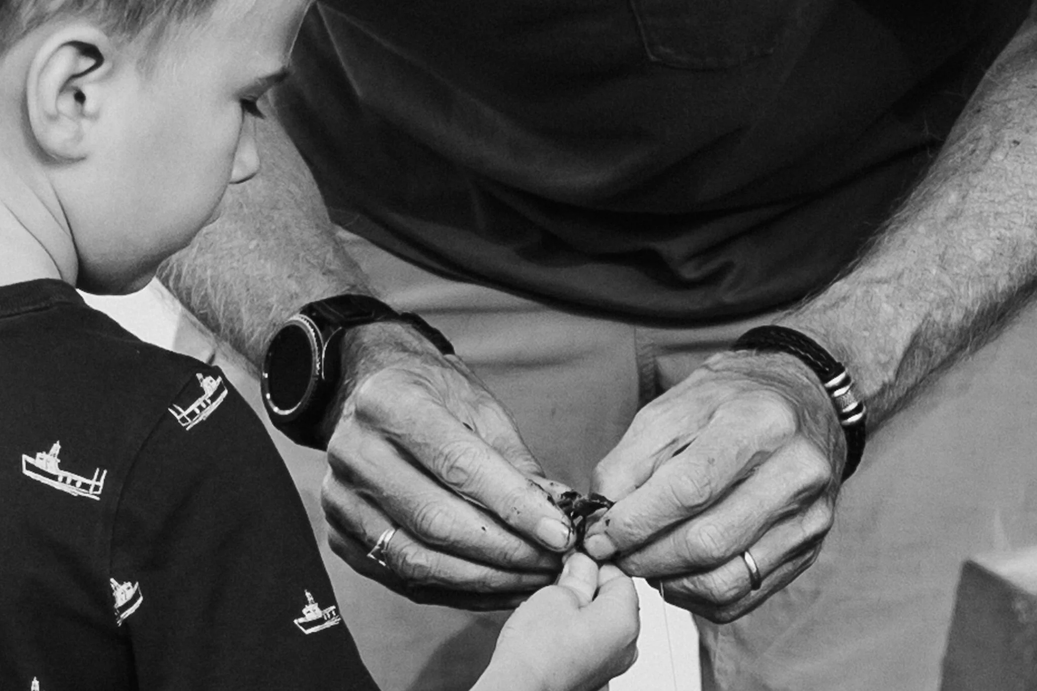 A young boy receives a vaccination from an adult man, whose hands are shown administering the shot. The boy is wearing a shirt with small sailboats, and the man is wearing a bracelet and a watch.