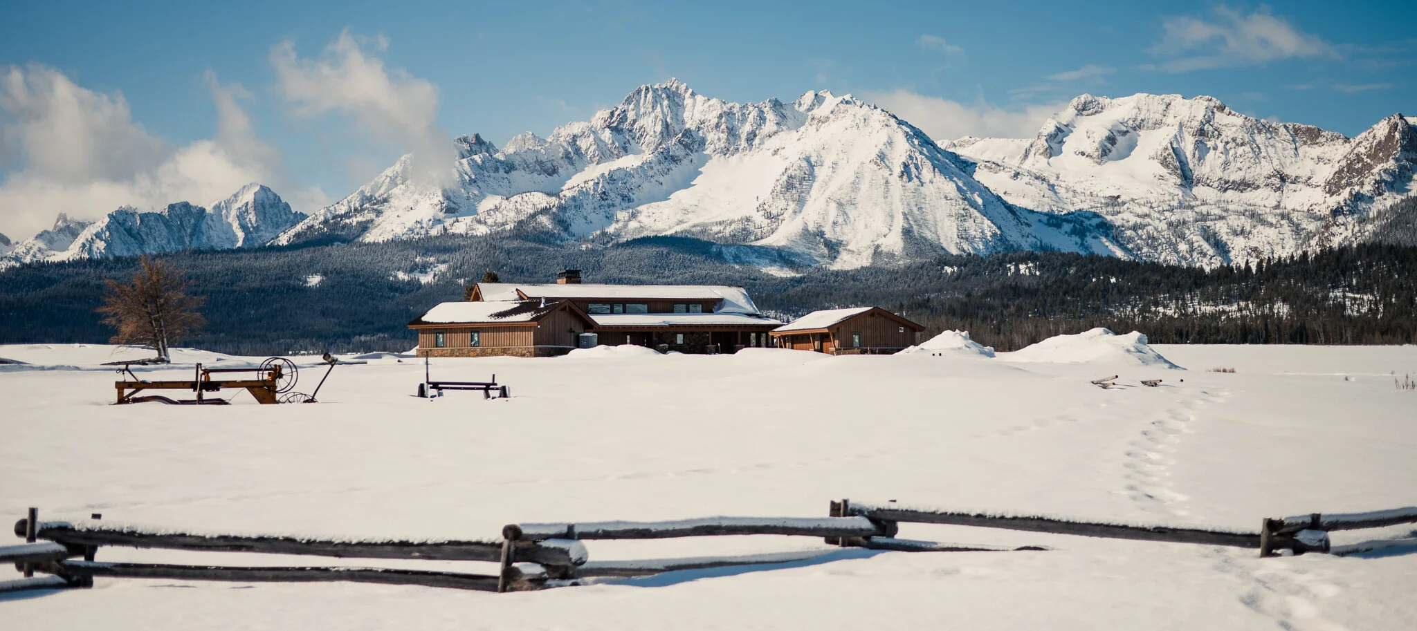 Snow-covered landscape with a mountain range in the background, a wooden house, and benches in the foreground.