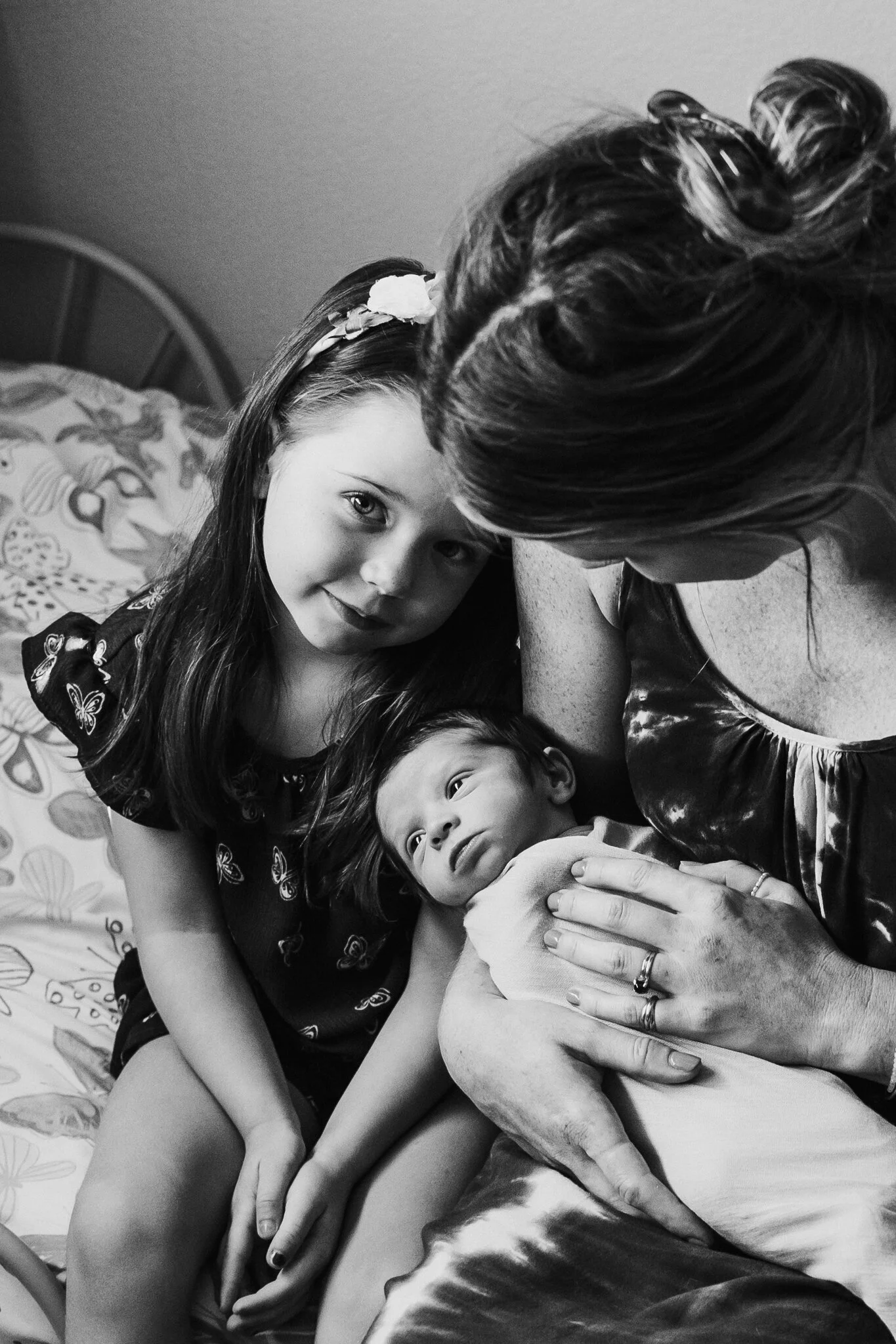 A woman with two rings holding a newborn baby, with a young girl beside her, all in a close, intimate moment on a bed.